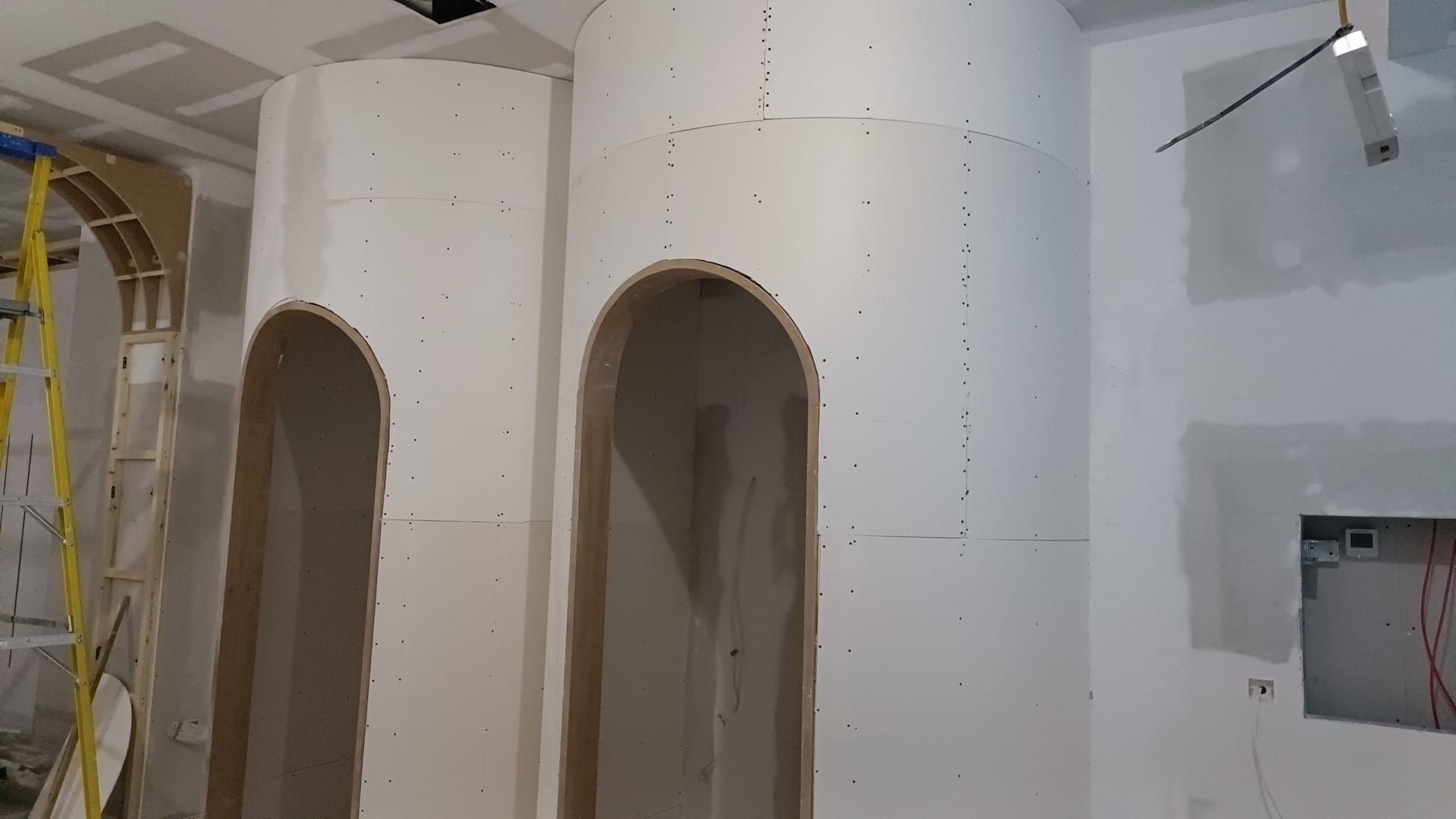 Interior construction site with white drywall columns and arched openings, unfinished walls, and a ladder on the left.