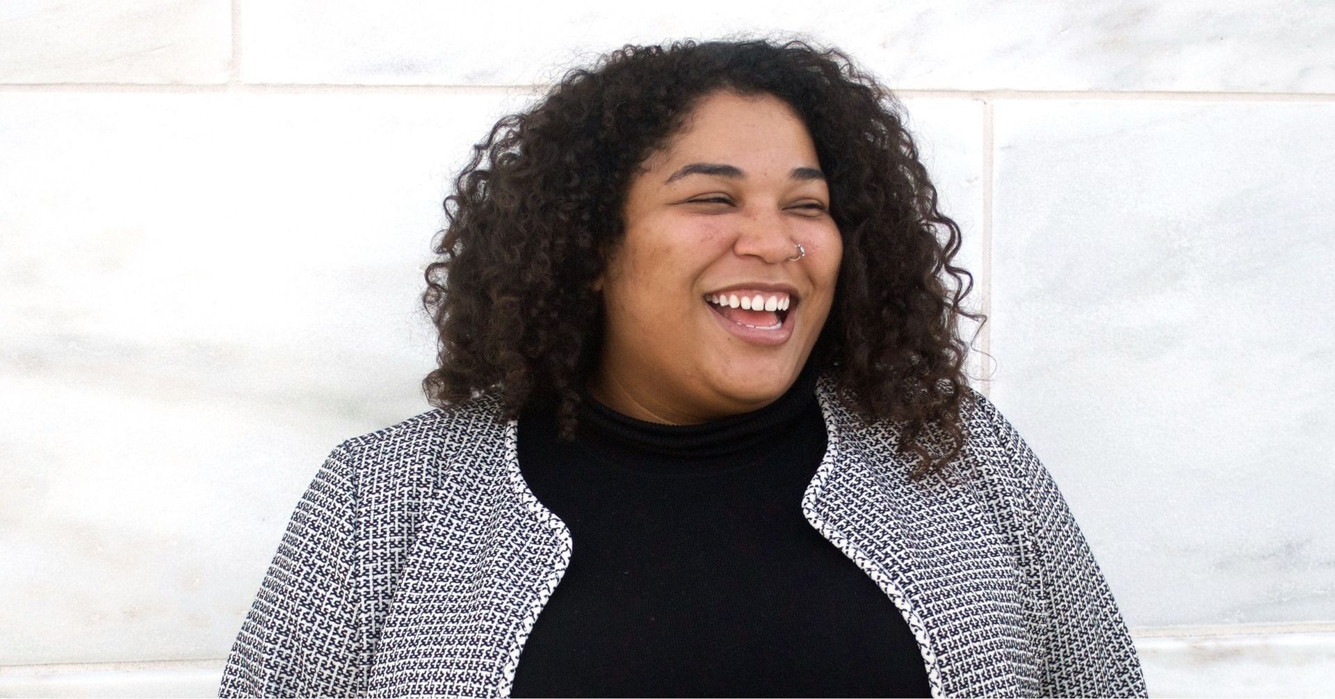 A woman with curly hair is laughing while standing in front of a white wall.