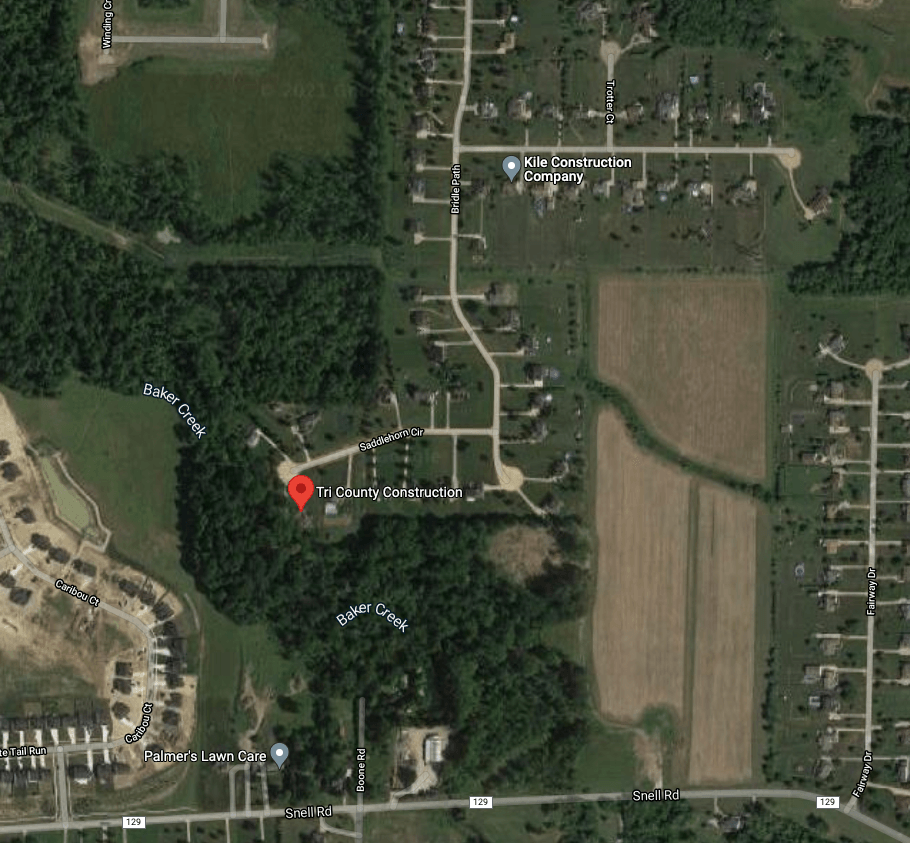 Aerial view of a suburban area with houses, roads, trees, and a field. Red location marker in wooded area.