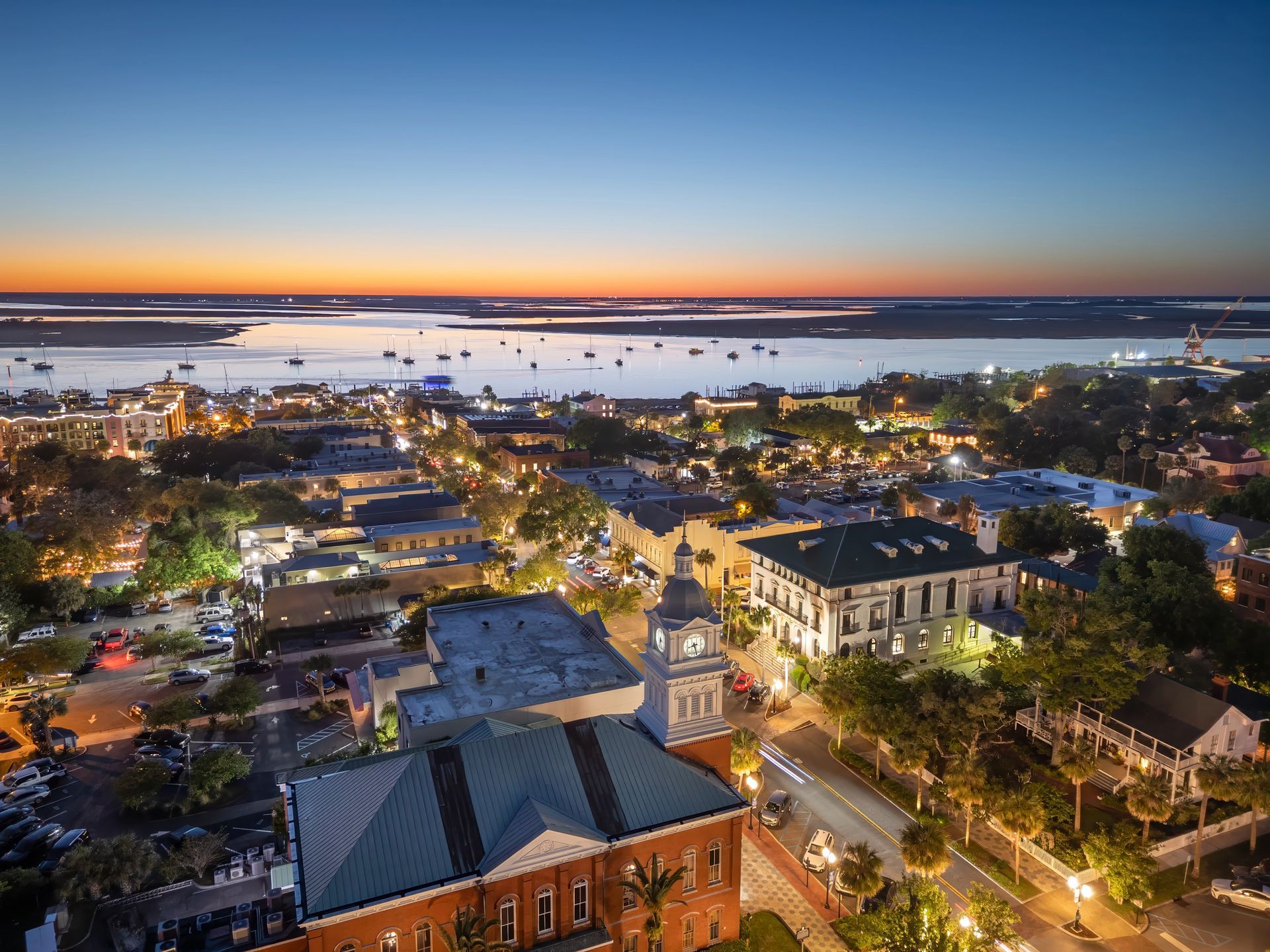 Aerial sunset view of a coastal town with lit streets, buildings, and boats on calm water.