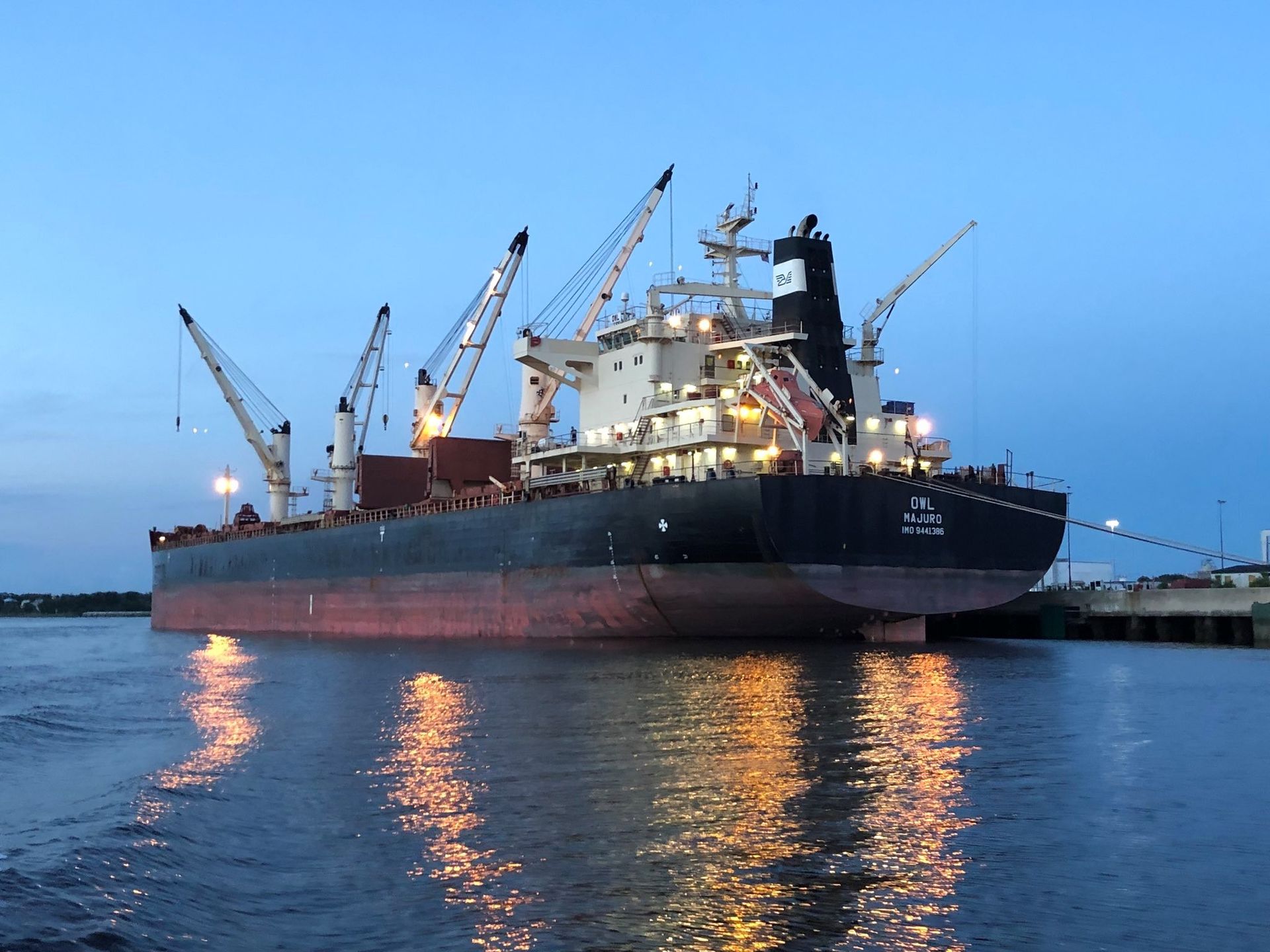 Cargo ship with cranes docked at sunset, reflecting on calm water.