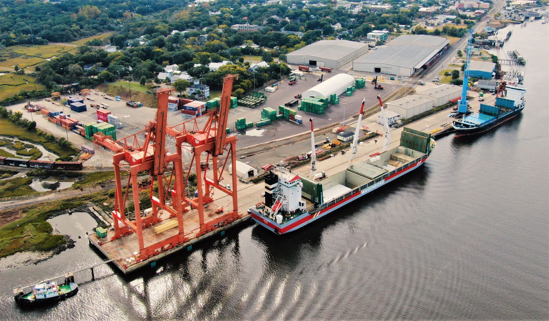 Port with orange cranes loading a cargo ship beside calm water and a dockside industrial yard