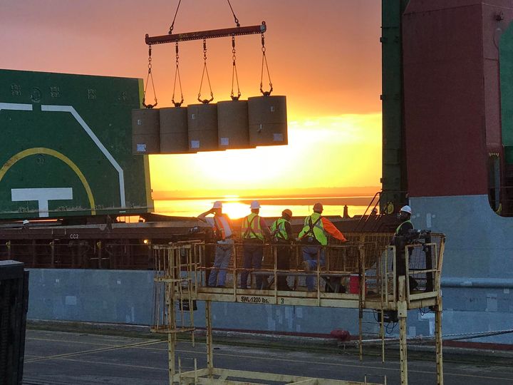 Workers on a platform unloading cargo at sunset beside a ship and crane.