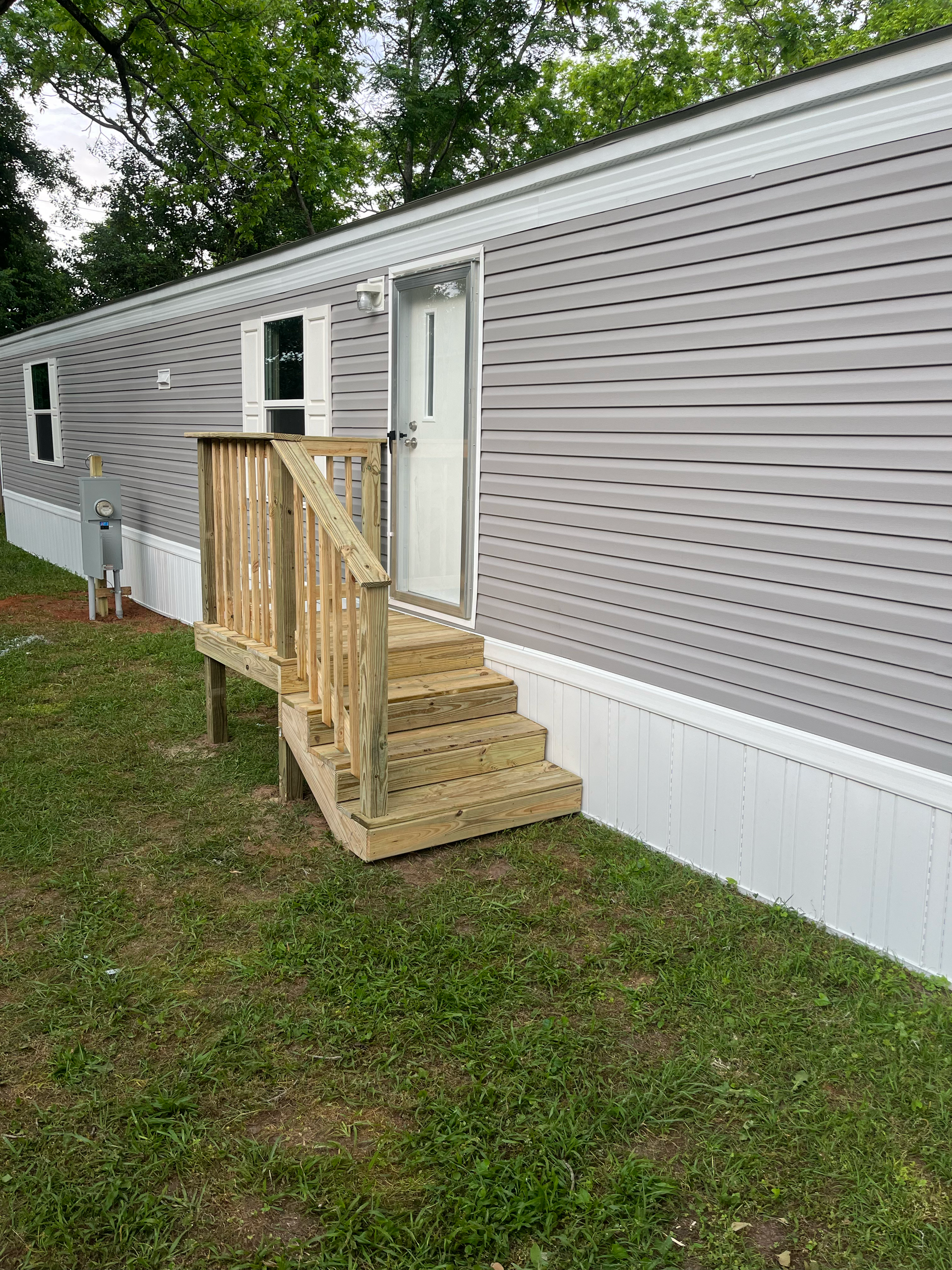 A gray mobile home with horizontal siding and a new, unfinished wooden staircase leading to a white door.