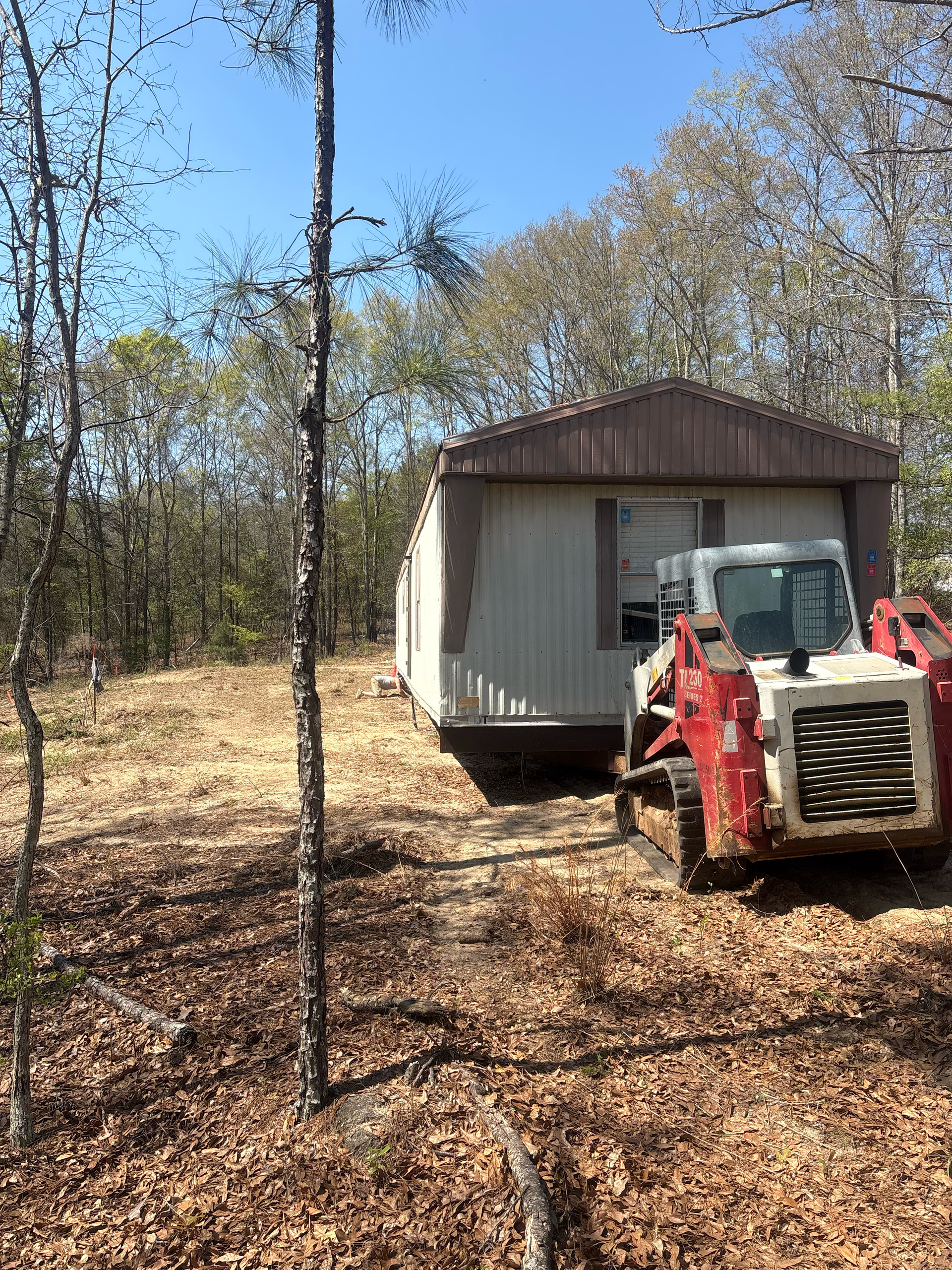A red skid steer loader parked next to a mobile home in a wooded area under a clear blue sky.