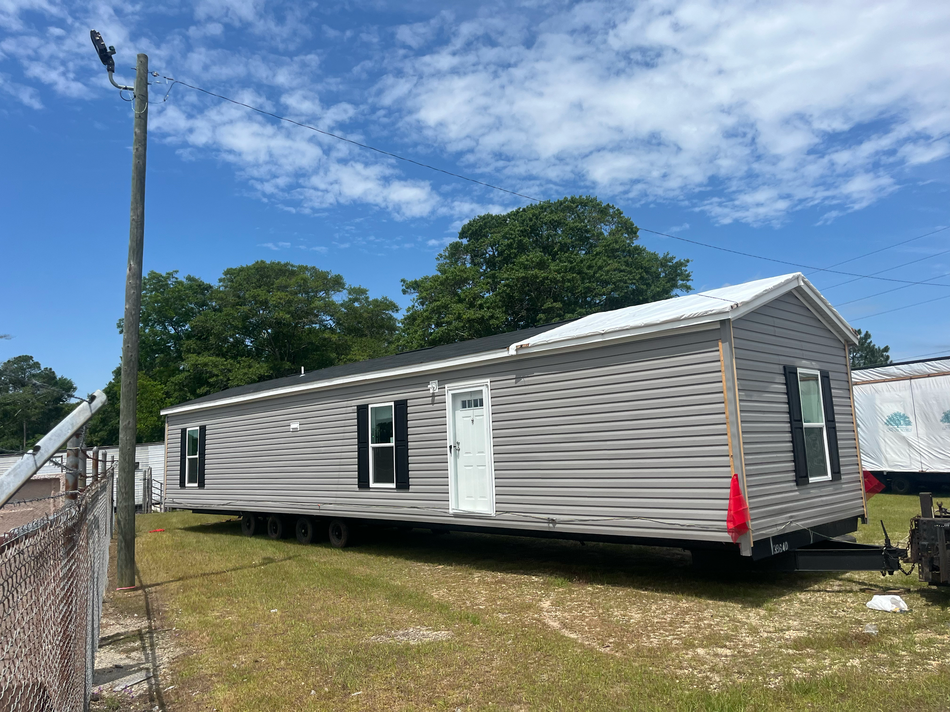 A grey, single-wide manufactured home on a metal frame, positioned on a grassy lot under a bright blue sky.