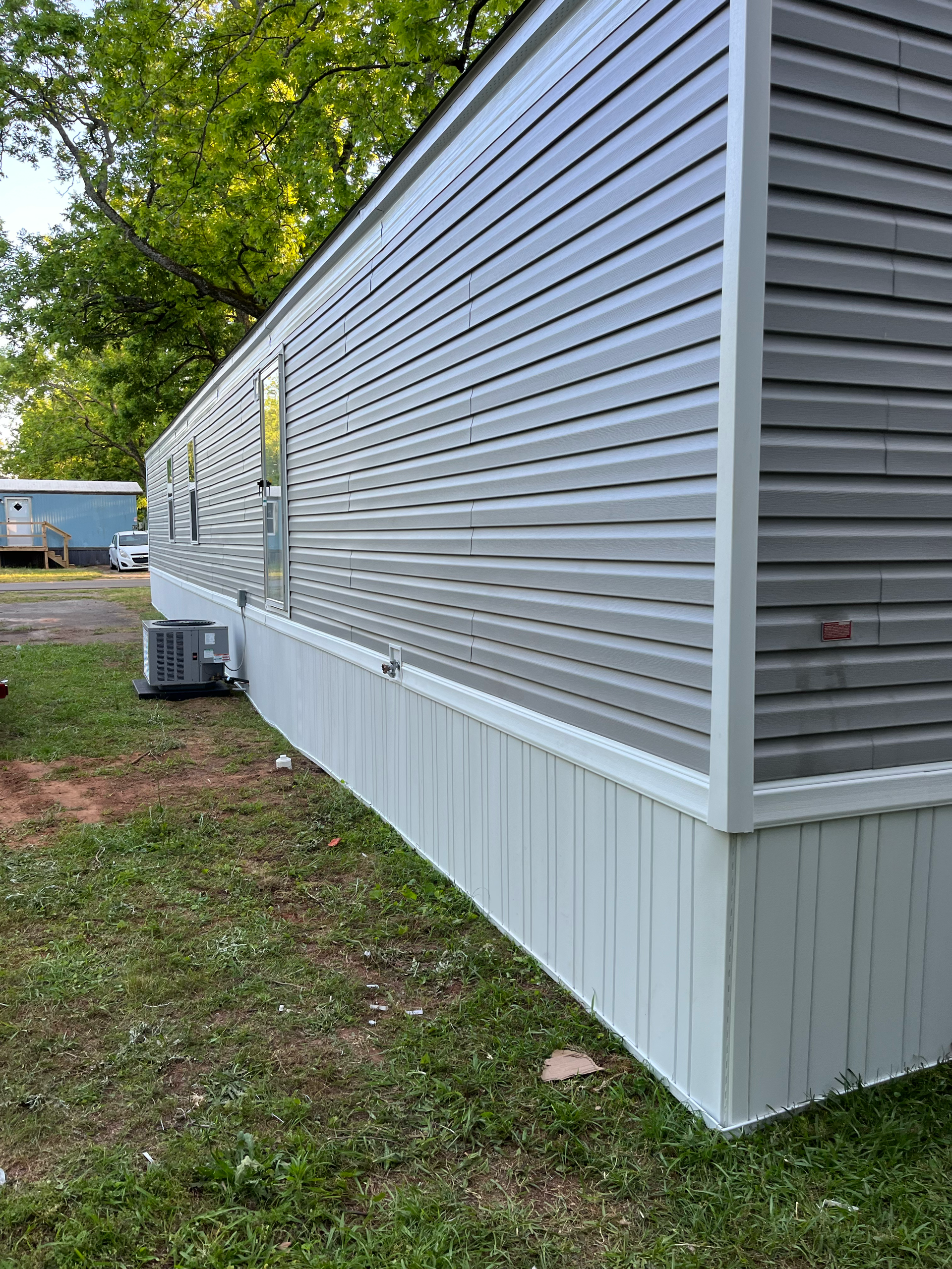 The side exterior of a grey mobile home with white skirting, set on a grass lawn with trees in the background.