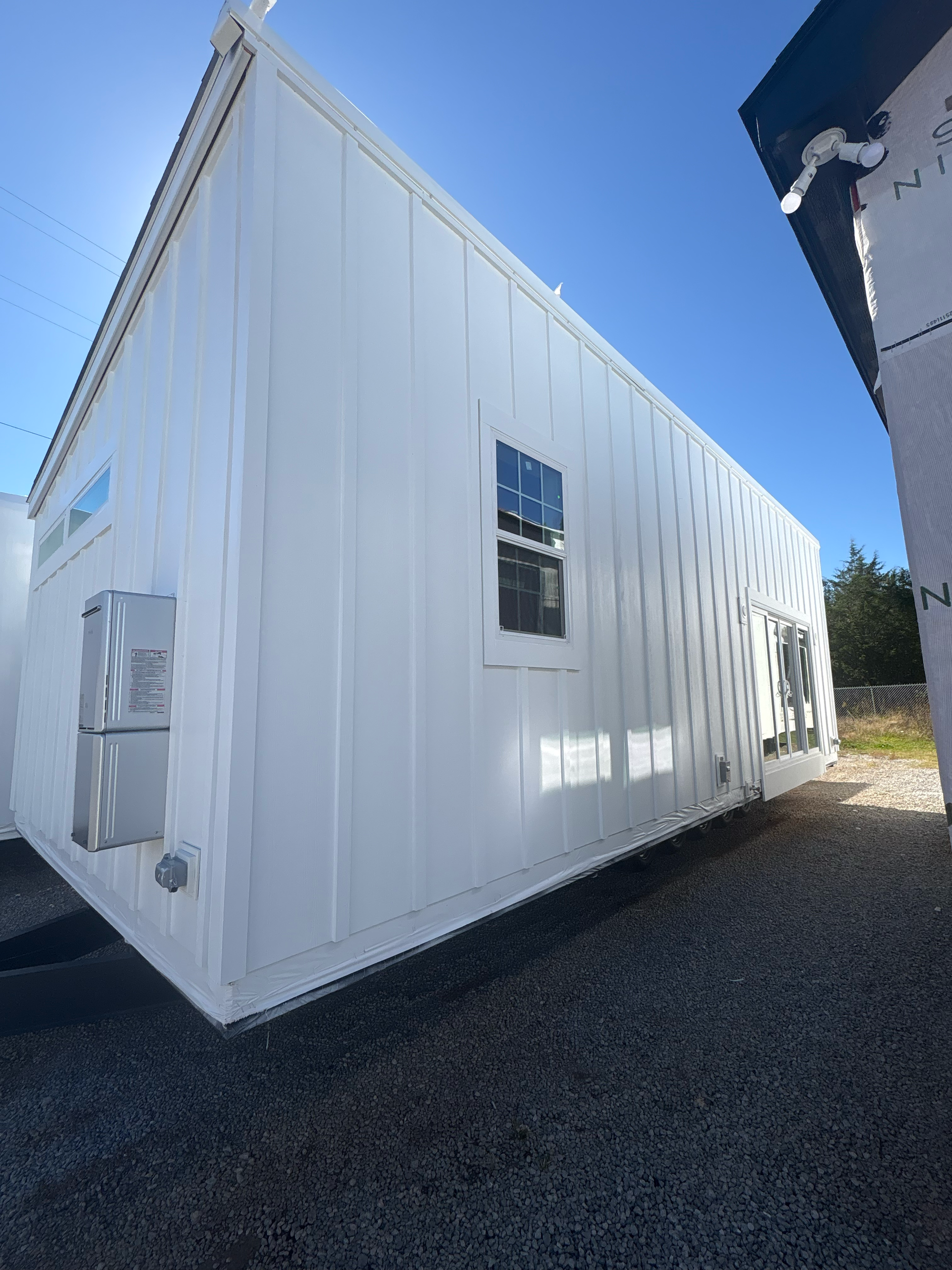 A white modular building with board-and-batten siding and one window stands on a gravel surface under a clear blue sky.