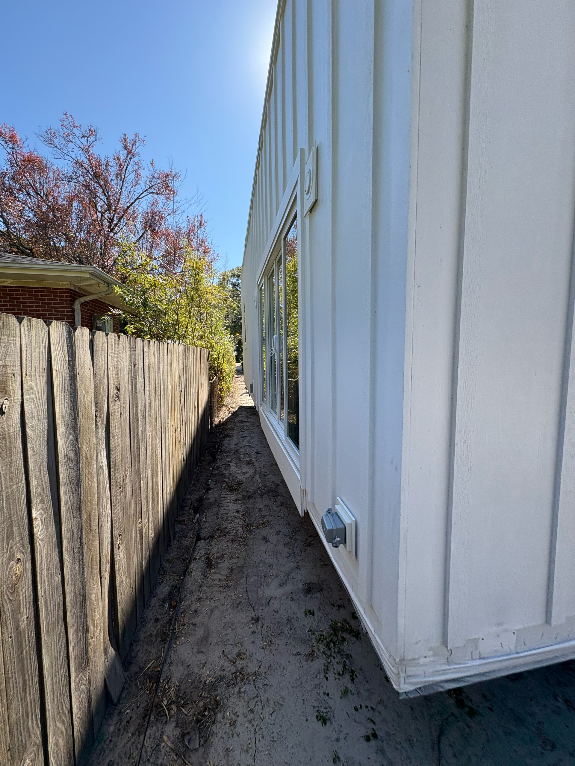 A narrow outdoor dirt path runs between a white building with vertical siding and a wooden fence under a clear blue sky.