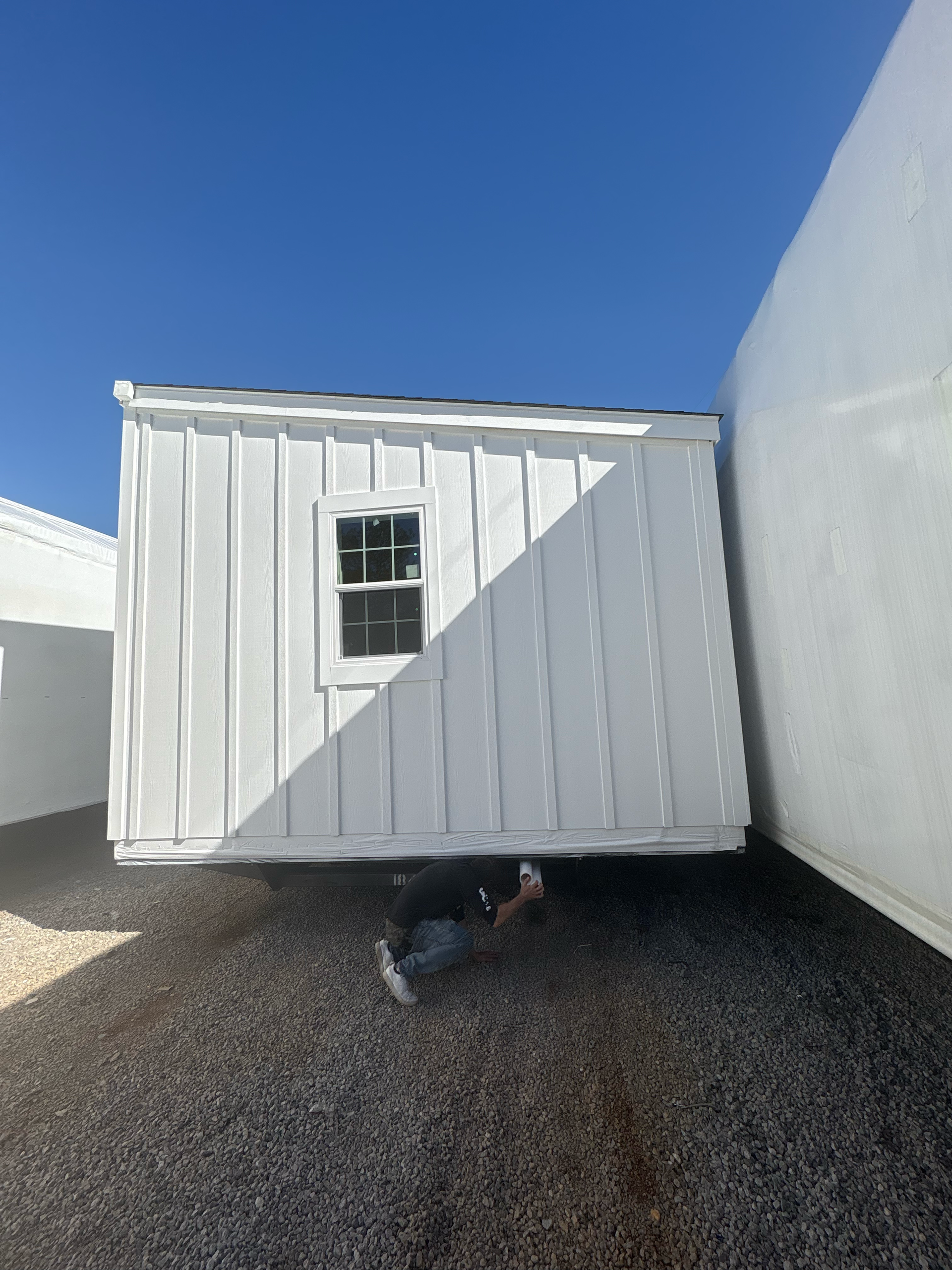 A person crouching under the raised edge of a white shed on a gravel lot under a clear blue sky.