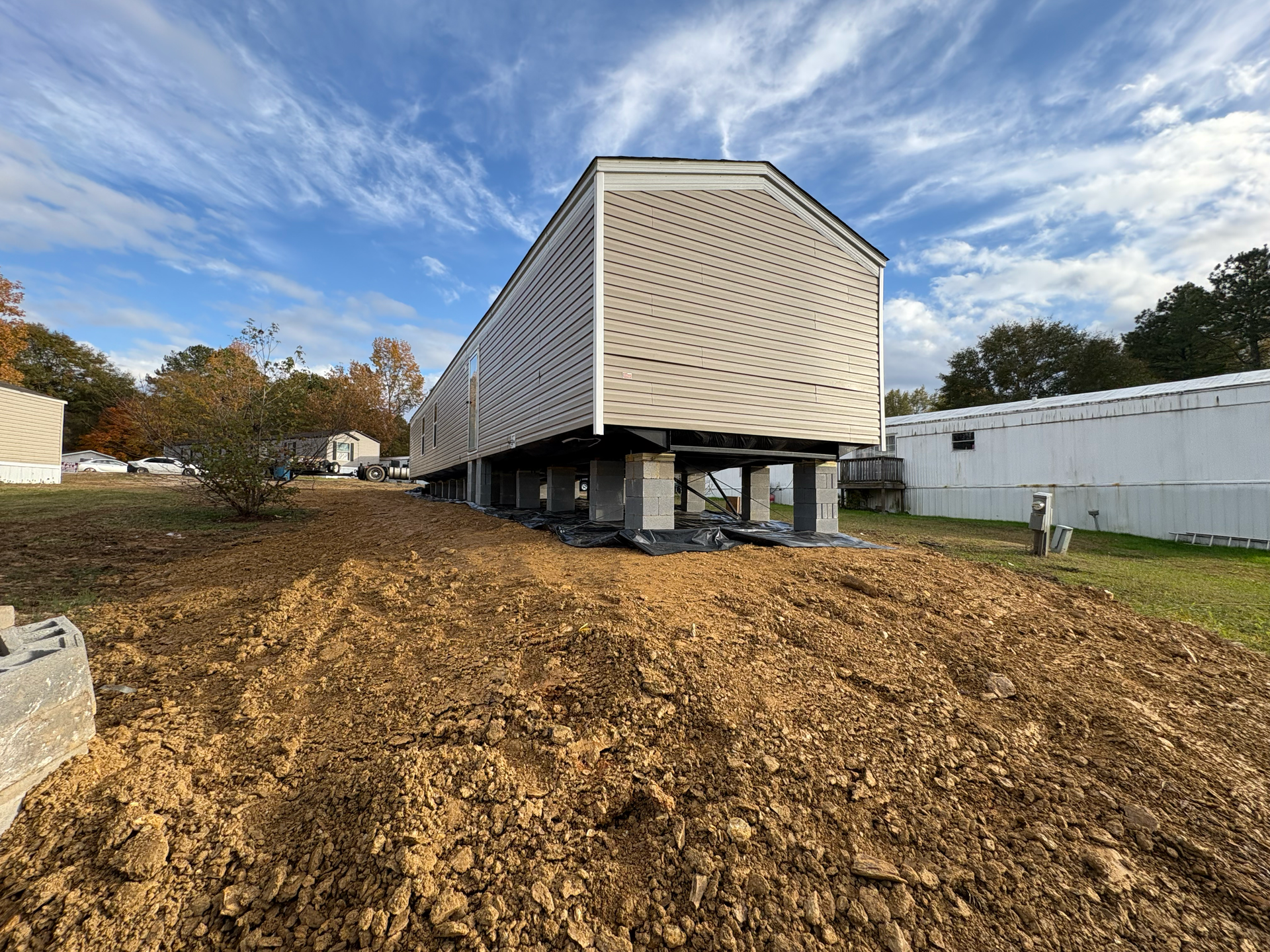 A manufactured home on cinder block pillars sits on a dirt lot under a cloudy blue sky.