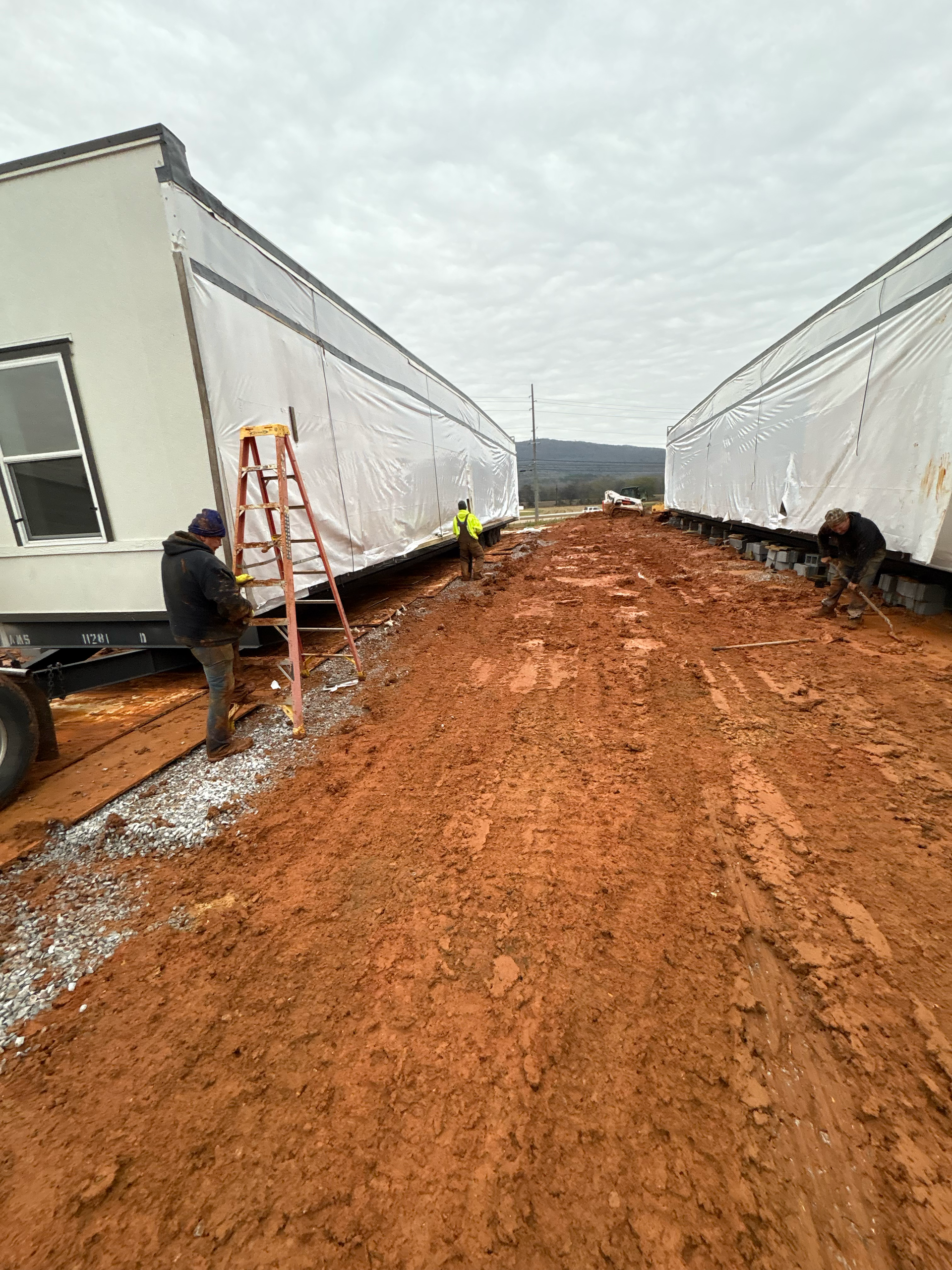Two workers work on modular home sections wrapped in plastic on a muddy, unpaved site under an overcast sky.