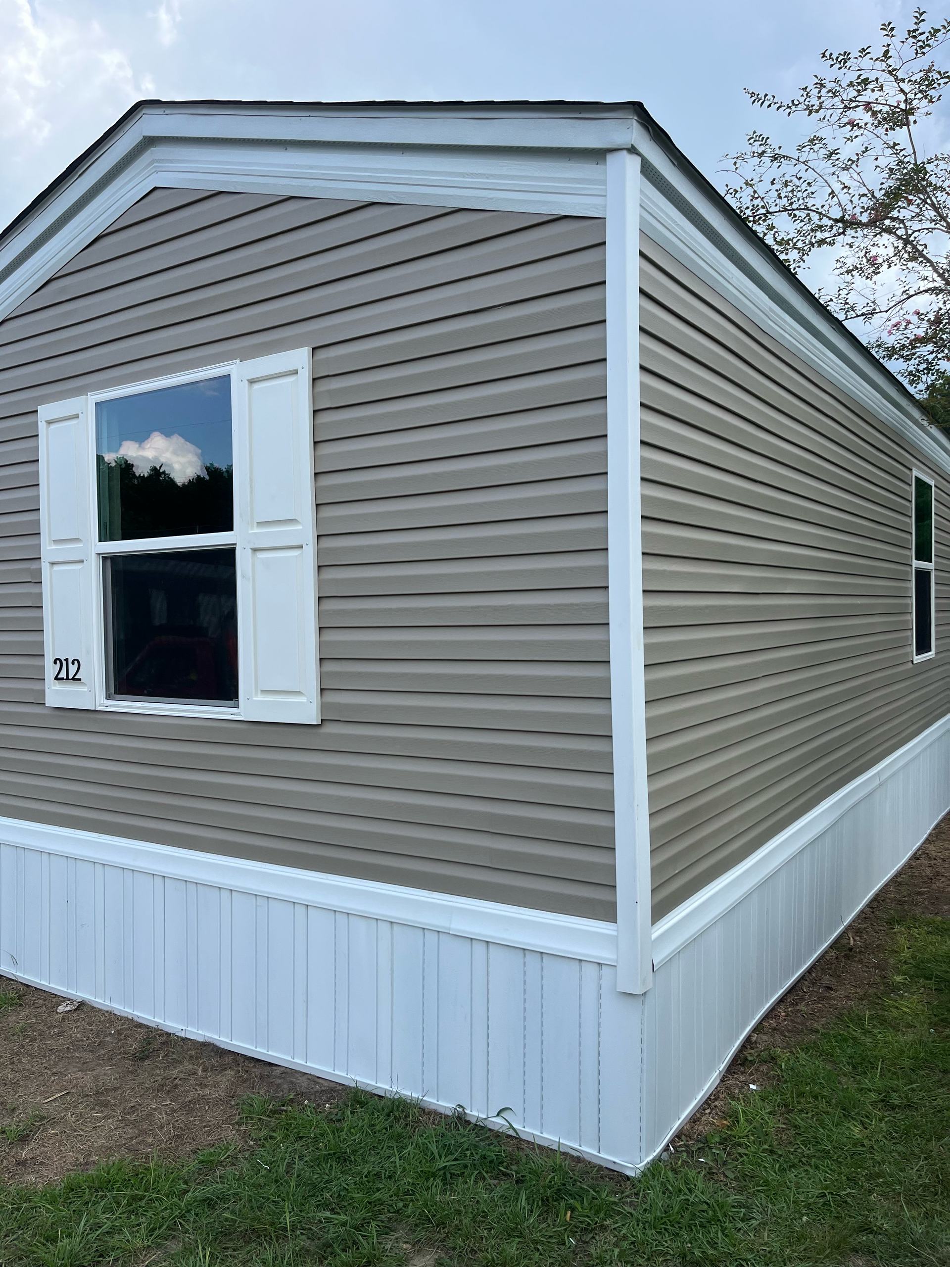 Exterior corner view of a manufactured home with light brown vinyl siding, white trim, white shutters, and white skirting.
