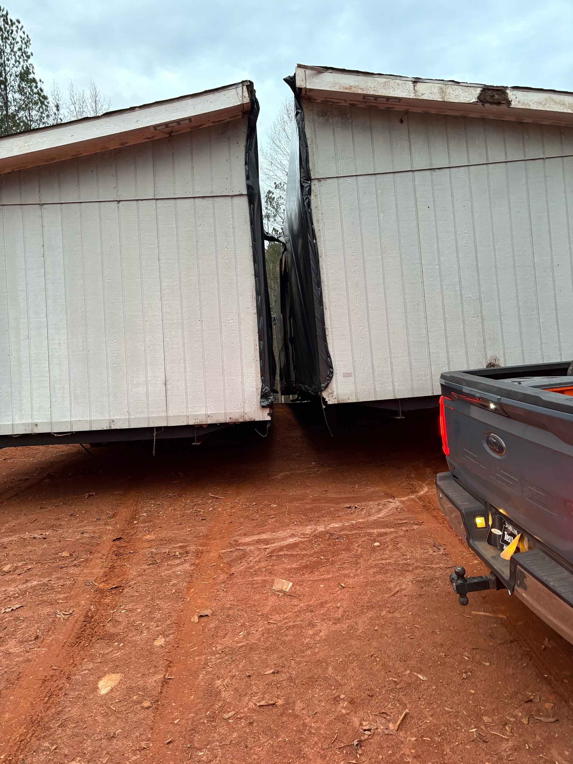 Two large white modular home sections sit side-by-side on red dirt near the back of a dark pickup truck.
