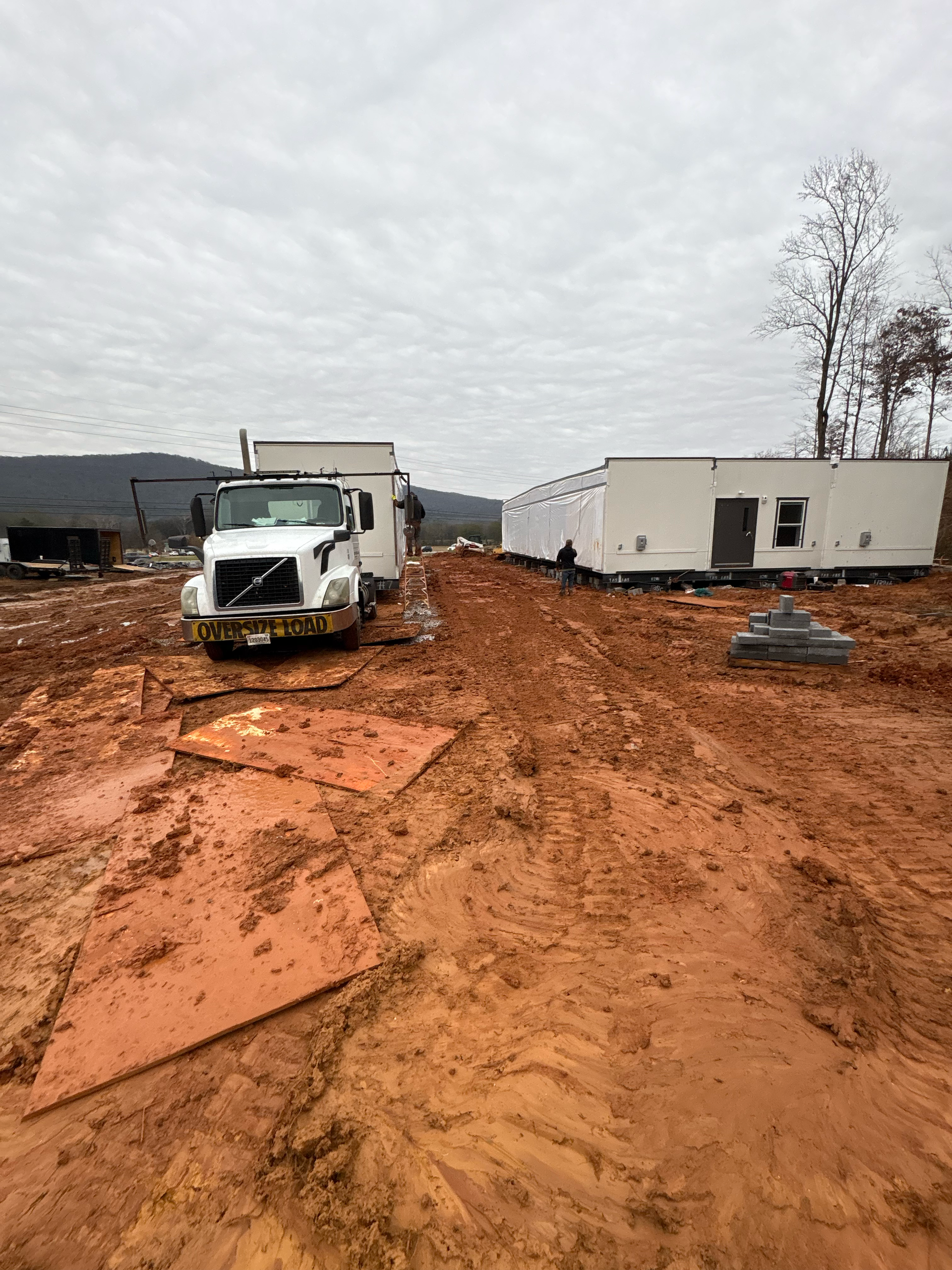 A white truck pulling a mobile unit on a muddy, construction-site terrain with a long building structure in the distance.