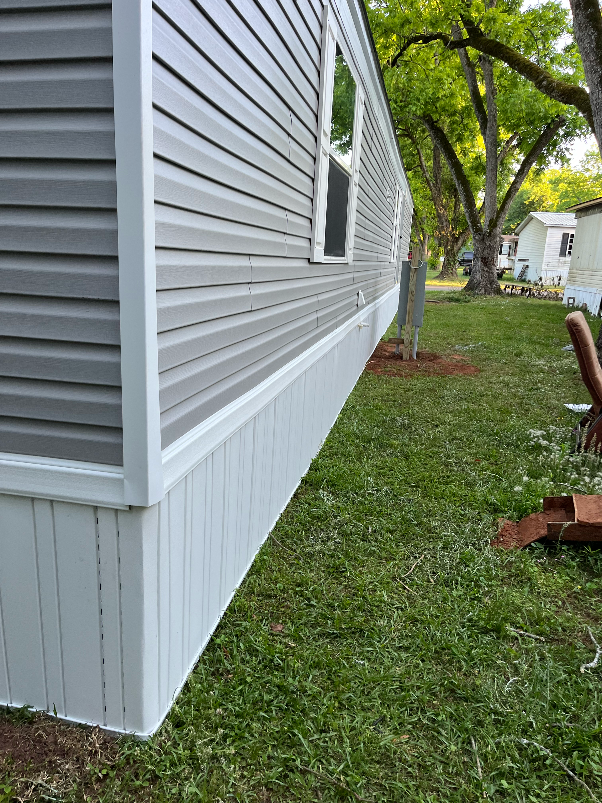 A view along the side of a manufactured home with gray horizontal siding and a white vertical skirt, set in a grassy yard.