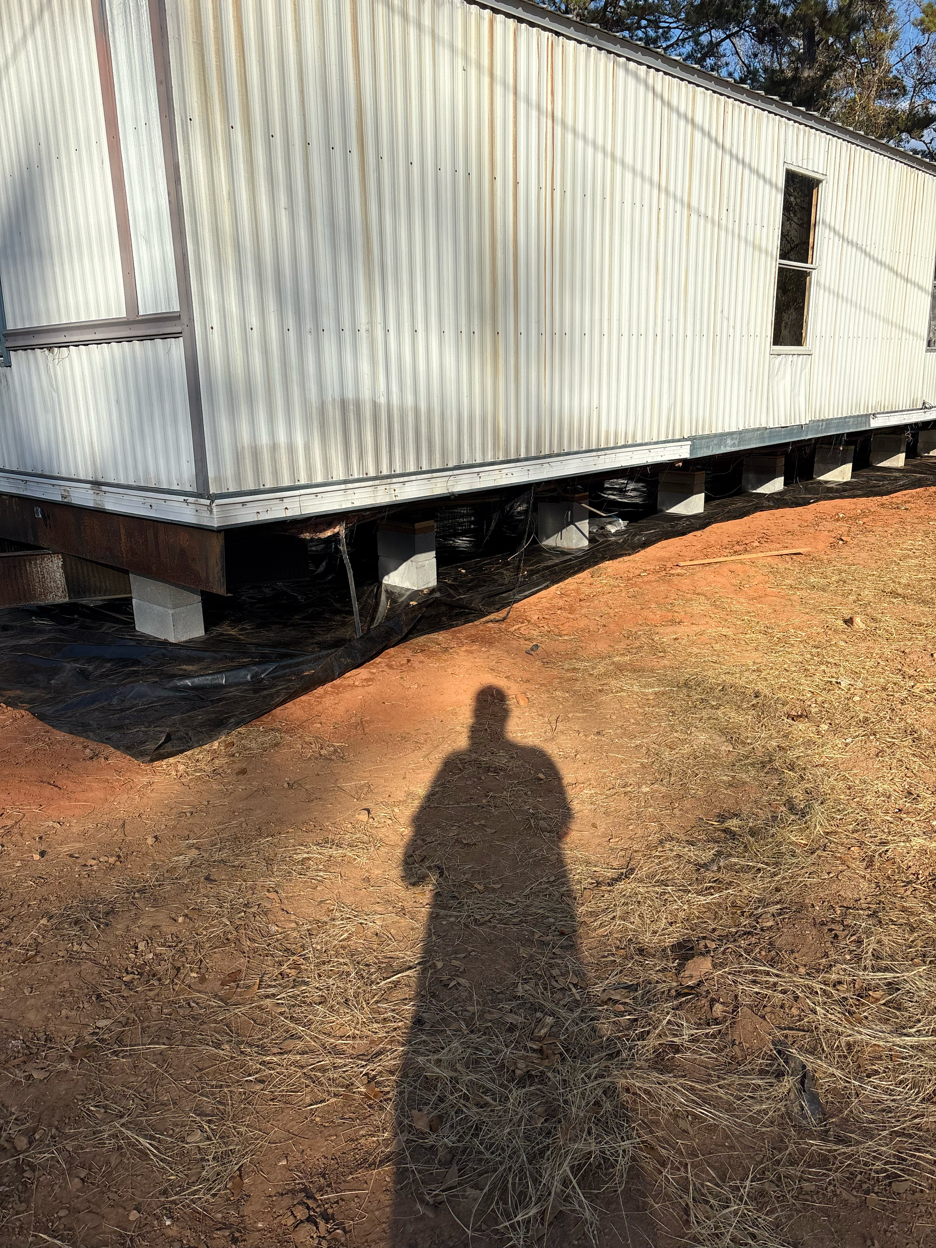 A long, white, corrugated metal mobile home on concrete block supports, with a person's shadow cast on dry grass.