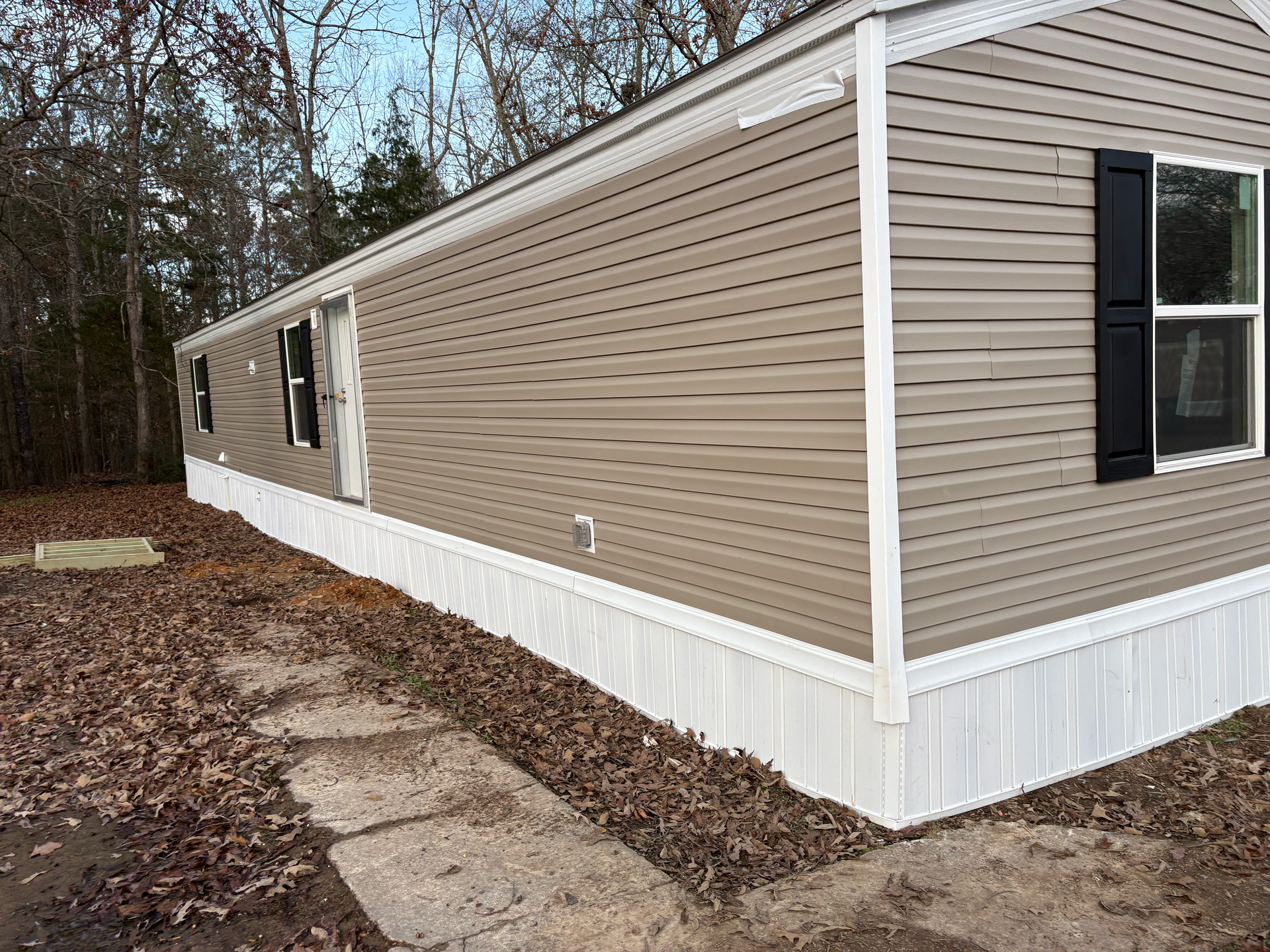 Tan-sided mobile home with white skirting and black window shutters, situated outdoors among fallen leaves and trees.