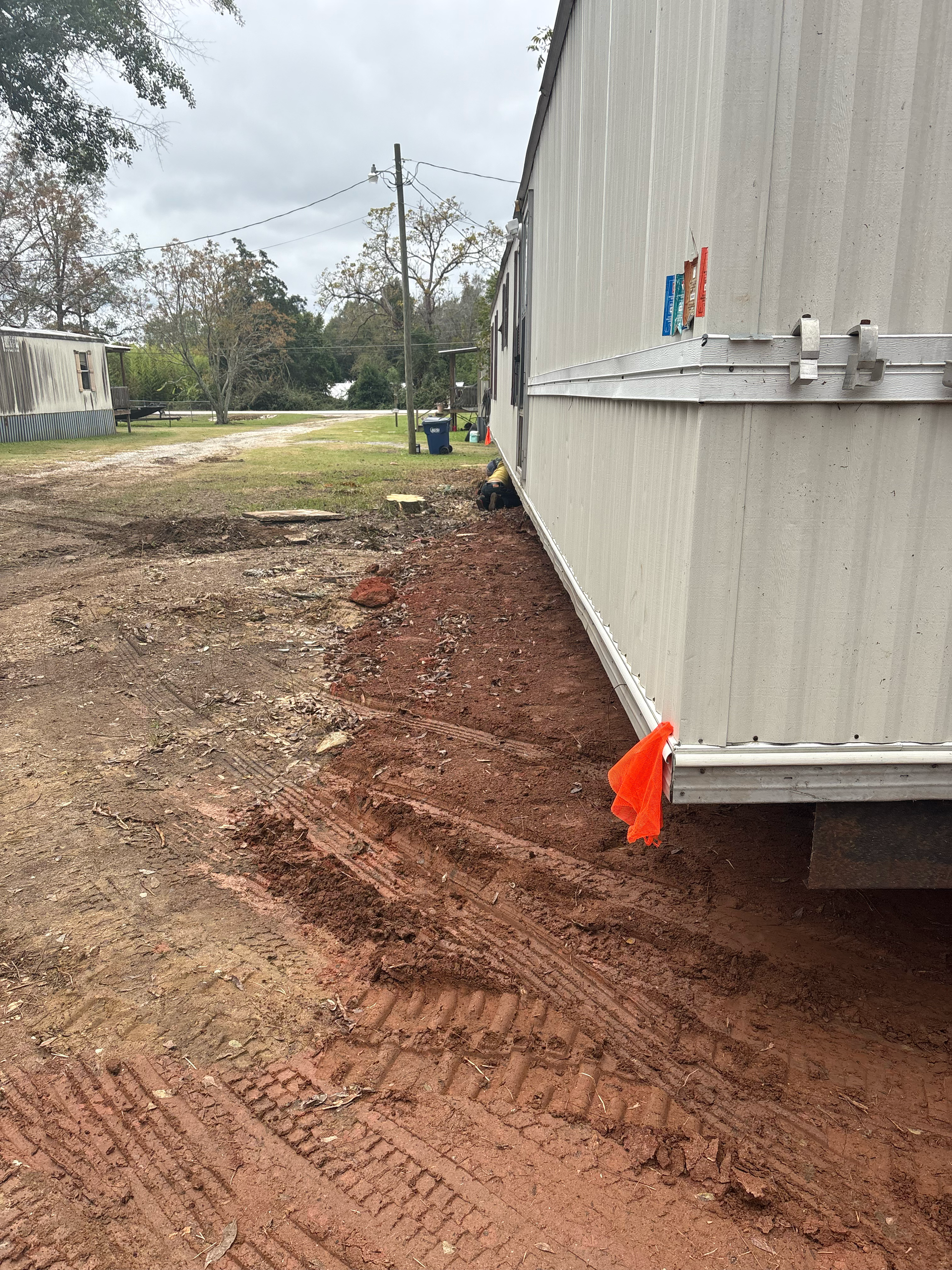 A side view of a light-colored mobile home on a dirt lot, featuring tire tracks in the red soil and an orange flag.