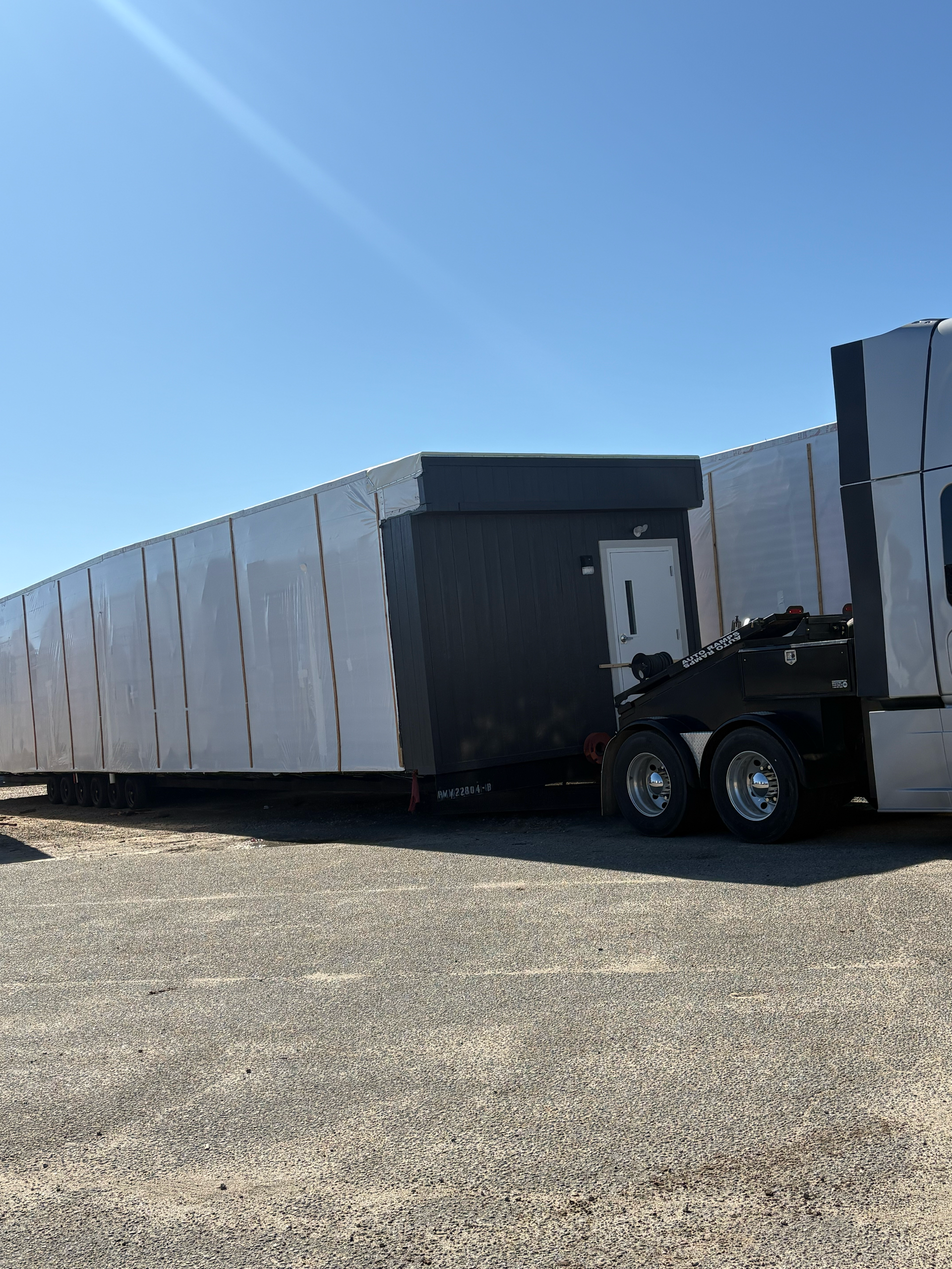 A semi-truck hauling a long, light-colored rectangular trailer with a dark-colored end section and a door.