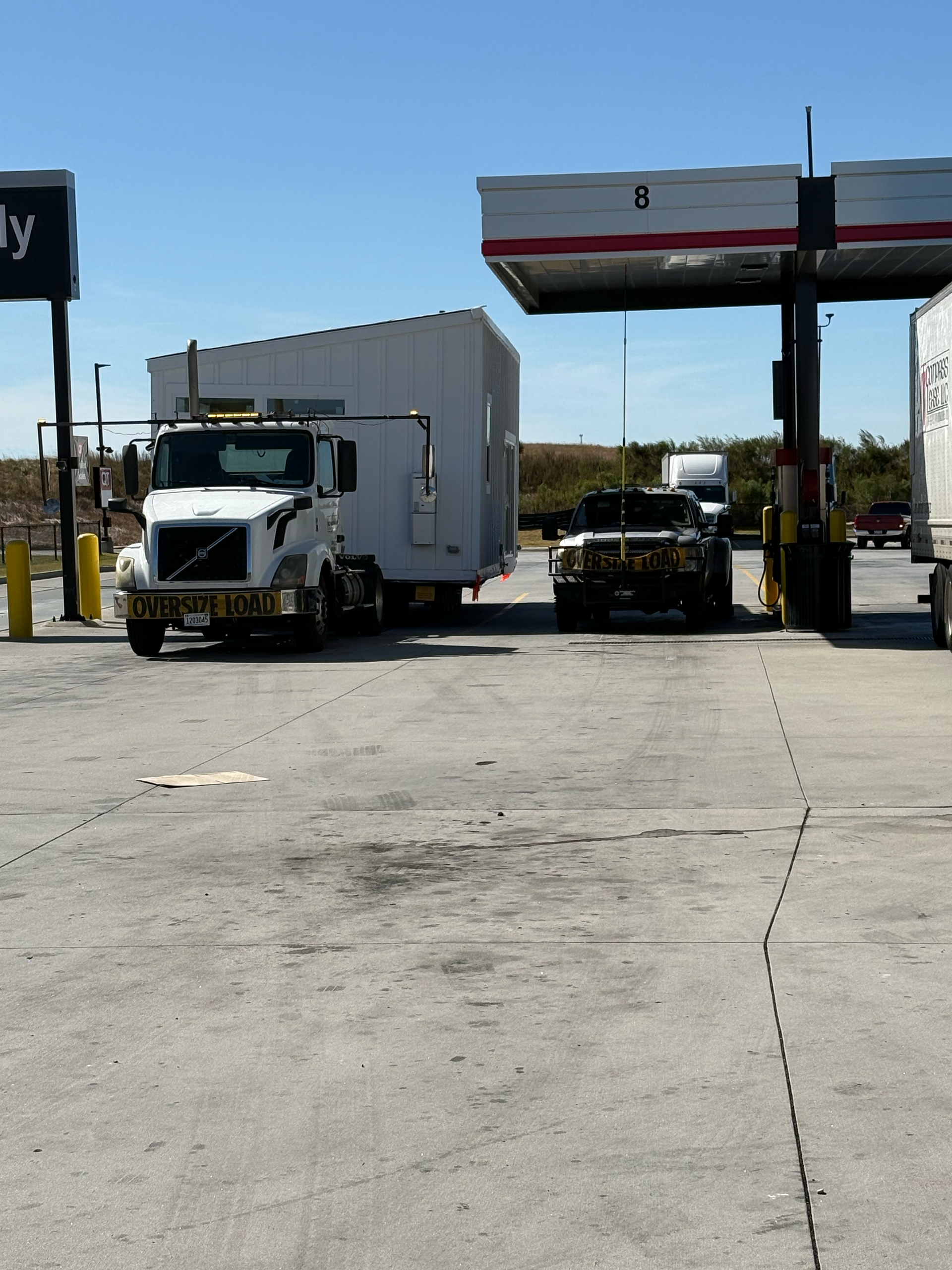 A white semi-truck hauling a large white trailer parked at a gas station under a canopy on a sunny day.