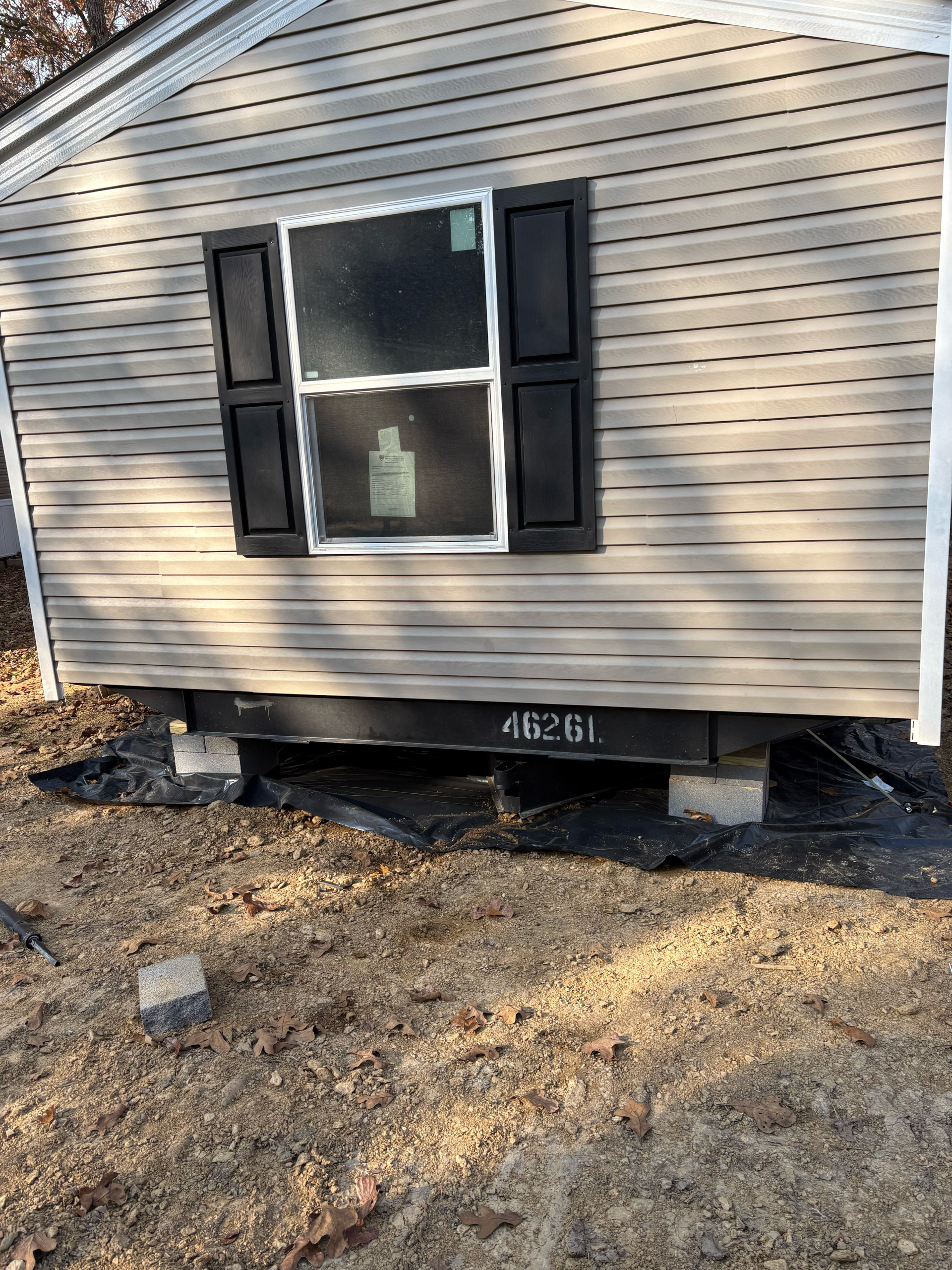 Tan vinyl-sided house section on concrete block pillars with a window and black shutters over a black ground tarp.