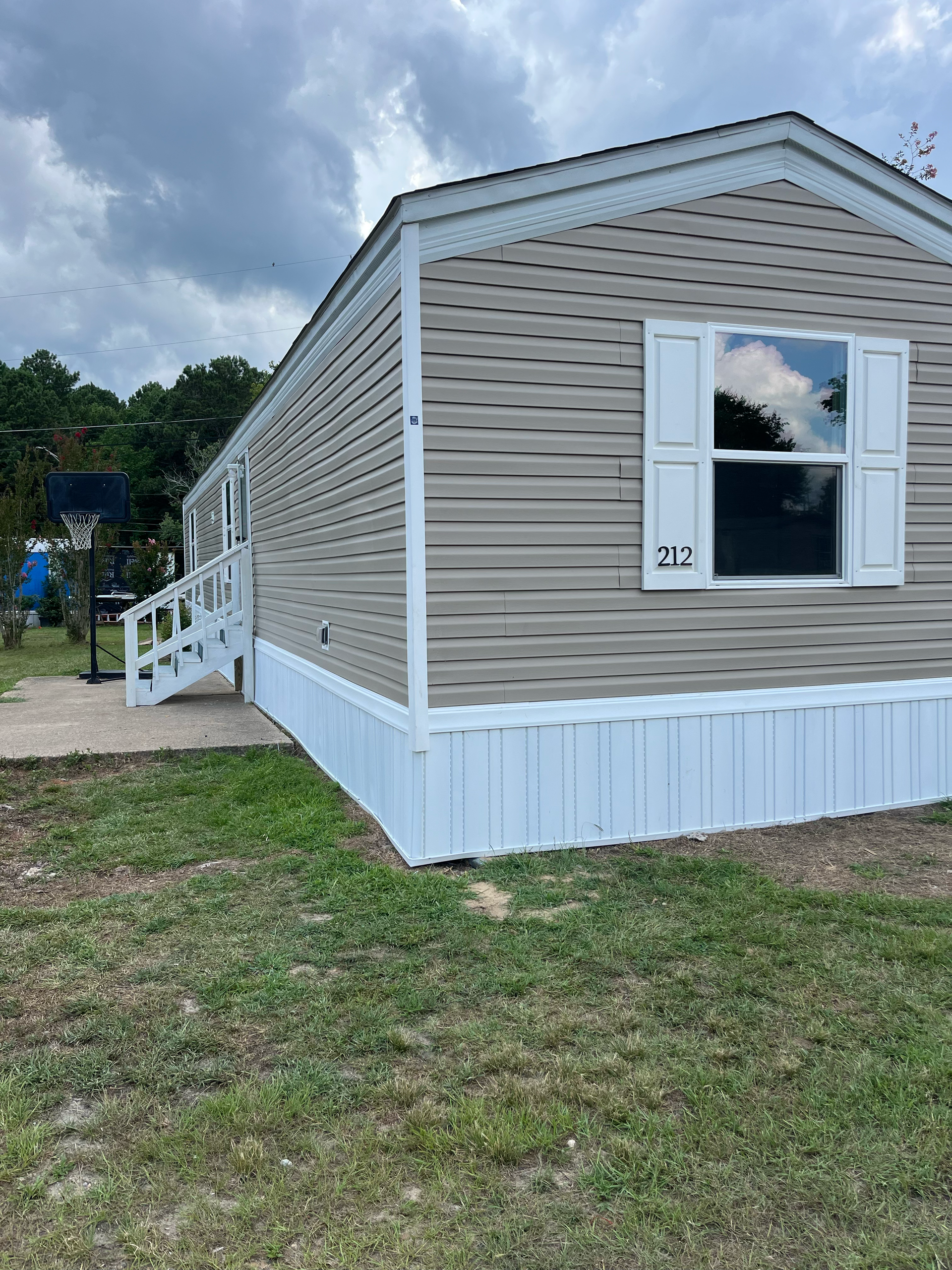 A tan mobile home with white skirting and stairs, a single window, and a basketball hoop in the yard.
