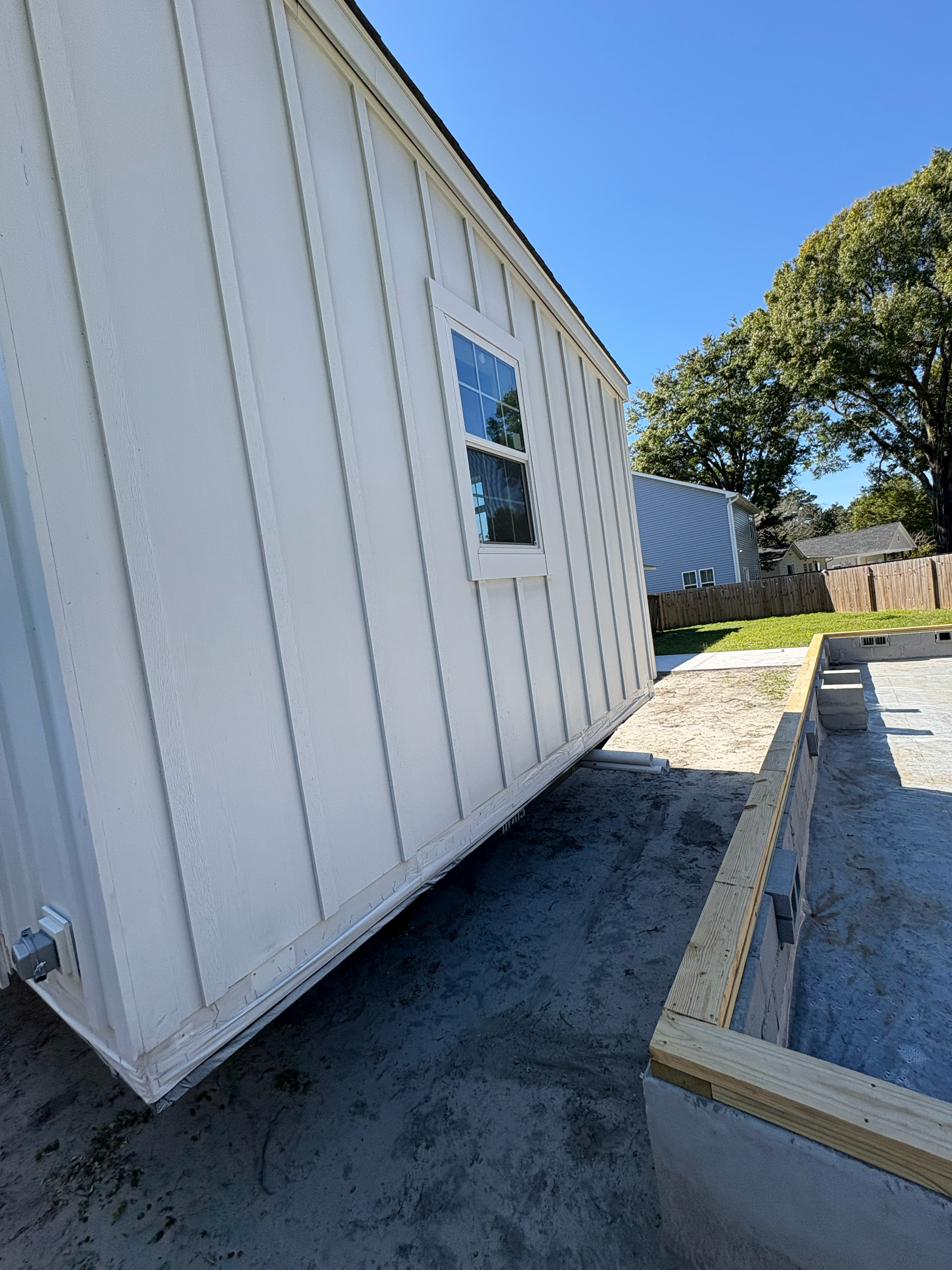 A white shed sits on a sandy lot next to a partially constructed wooden foundation with a concrete border.