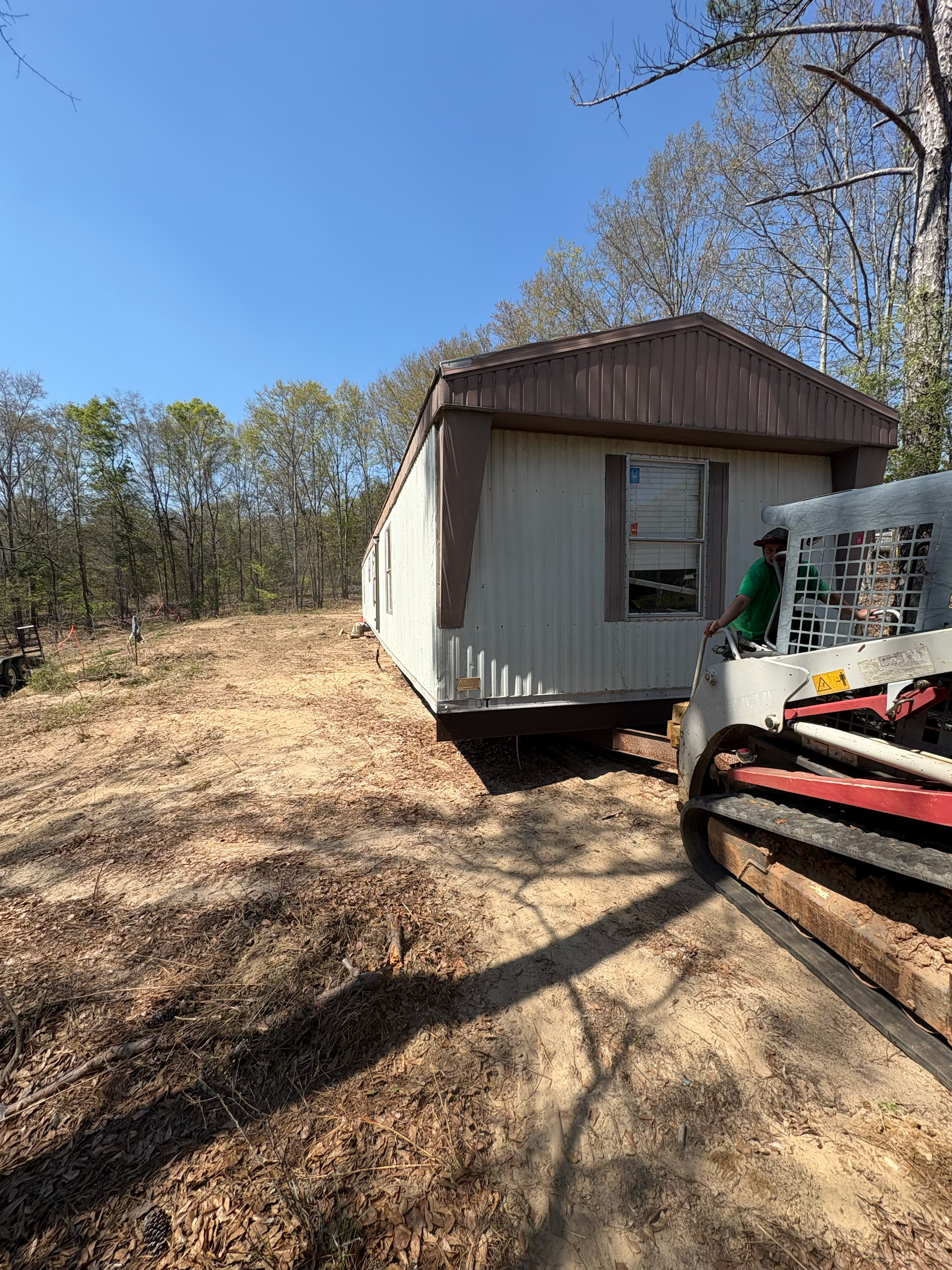 A worker operates a compact track loader to move a mobile home across a wooded, dirt-covered lot.