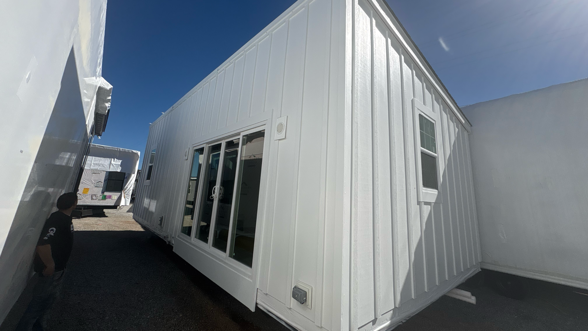 A white, modern, shed-style structure with large glass doors and vertical siding, parked in an outdoor gravel lot.