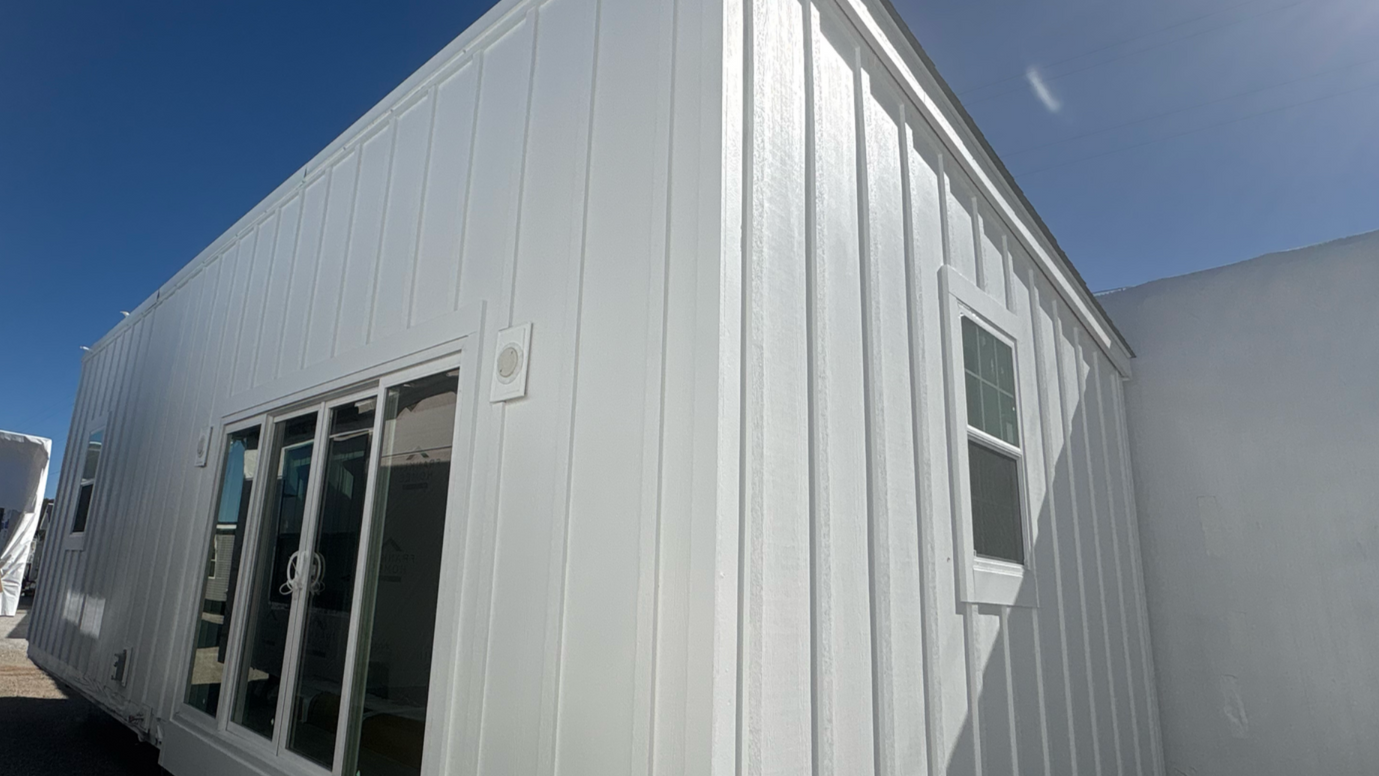 White wooden tiny home building exterior with glass doors and windows under a blue sky.