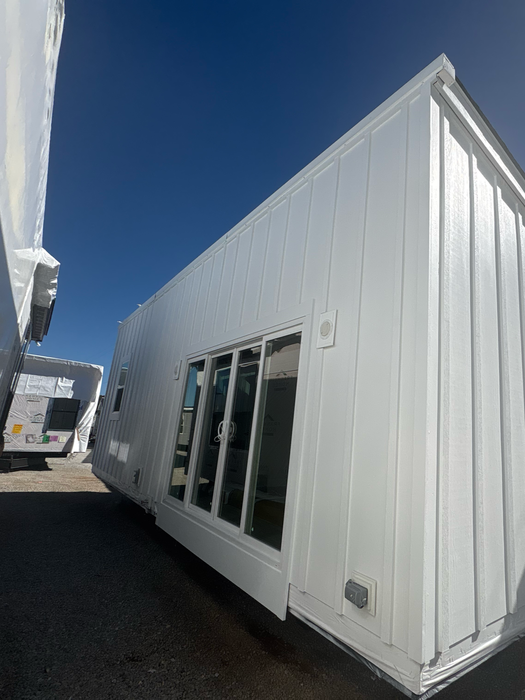 A modern, white tiny house with vertical board-and-batten siding and a glass sliding door, parked on a gravel lot.
