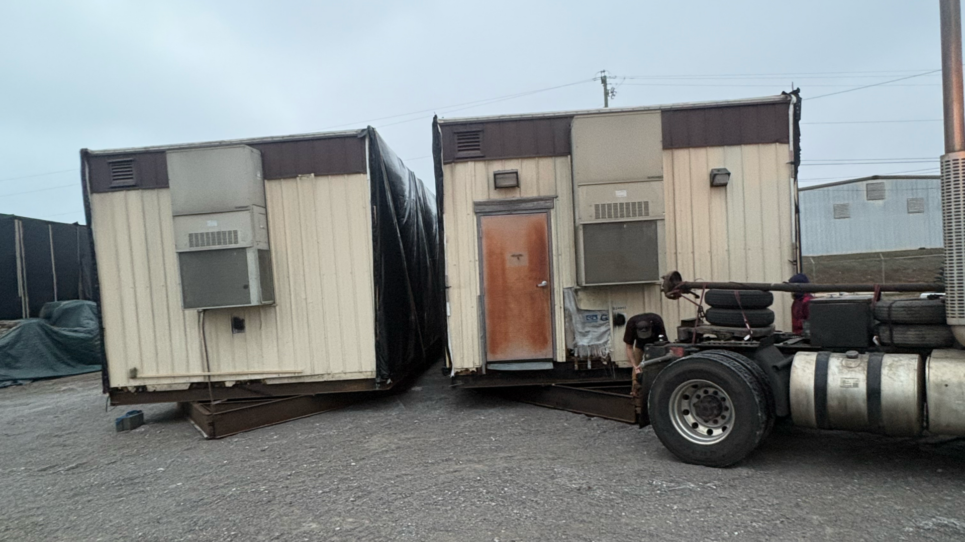 Two rectangular portable office trailers with beige and brown siding sit on a gravel lot next to a parked semi-truck.