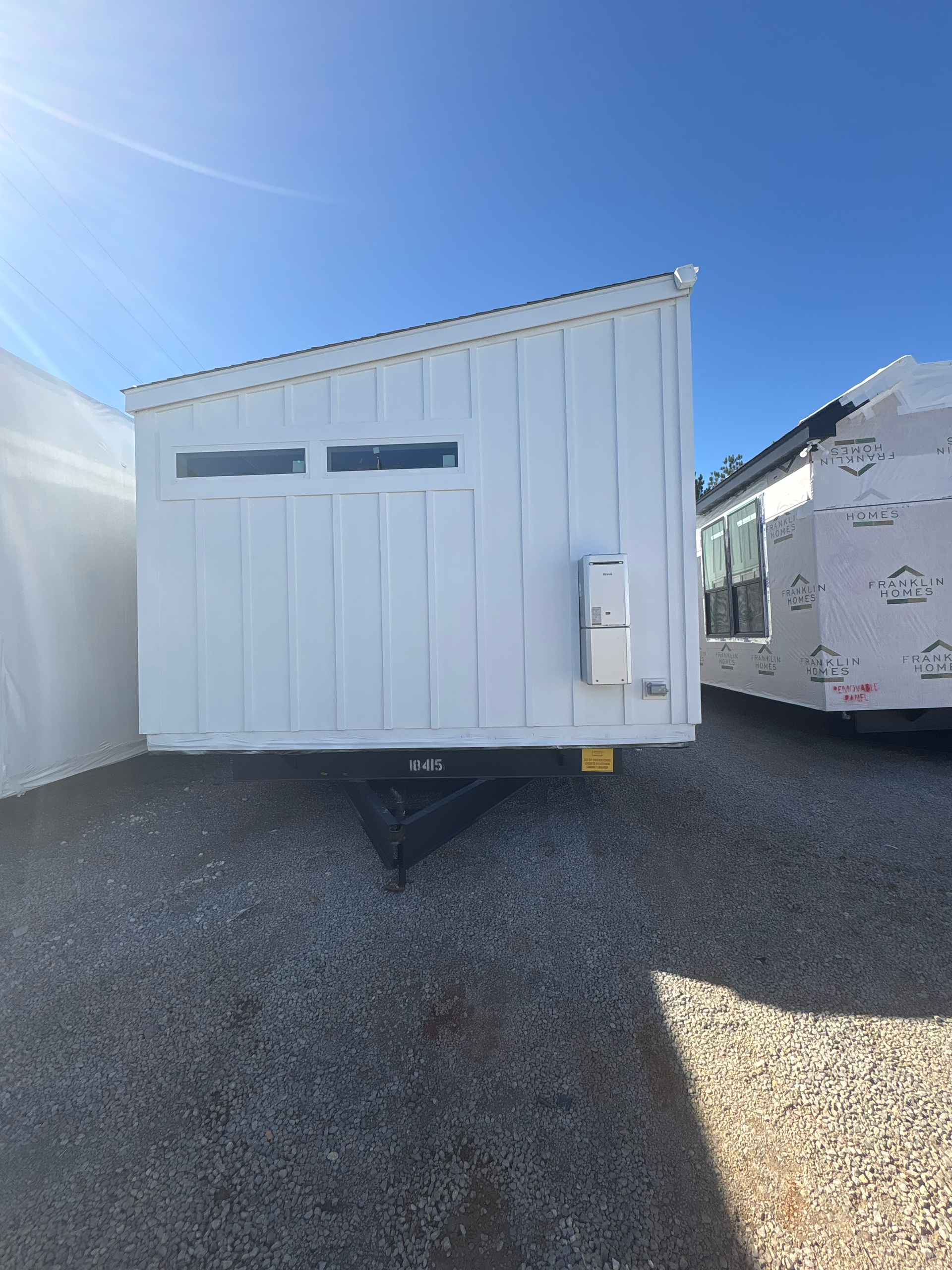 A white, modern tiny home with a shed roof and two horizontal windows parked on a gravel lot under a clear blue sky.