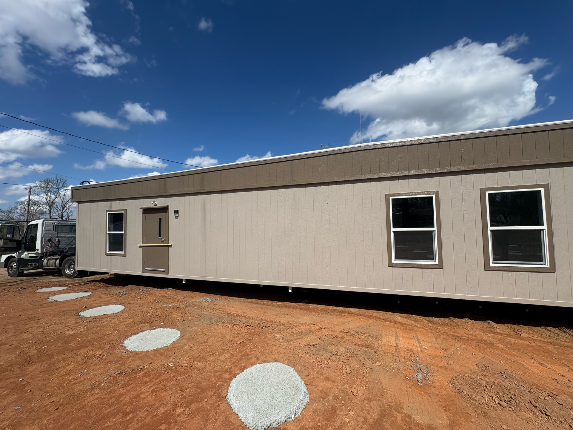 Tan modular building on a red dirt lot under a sunny blue sky with cloud-shaped stepping stones leading to the door.