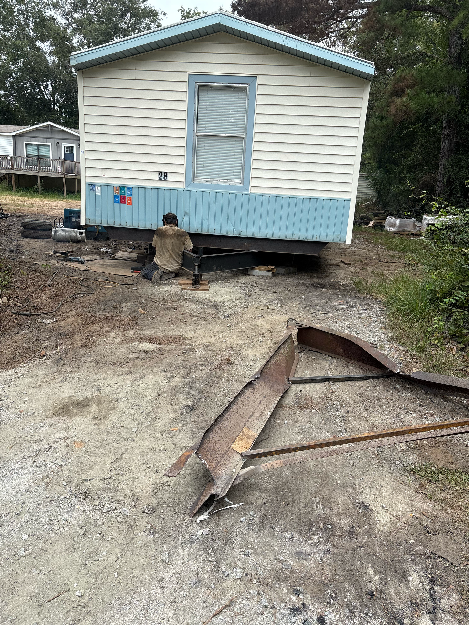A person works underneath a light-colored mobile home, with a detached, rusted metal trailer tongue lying on the ground.