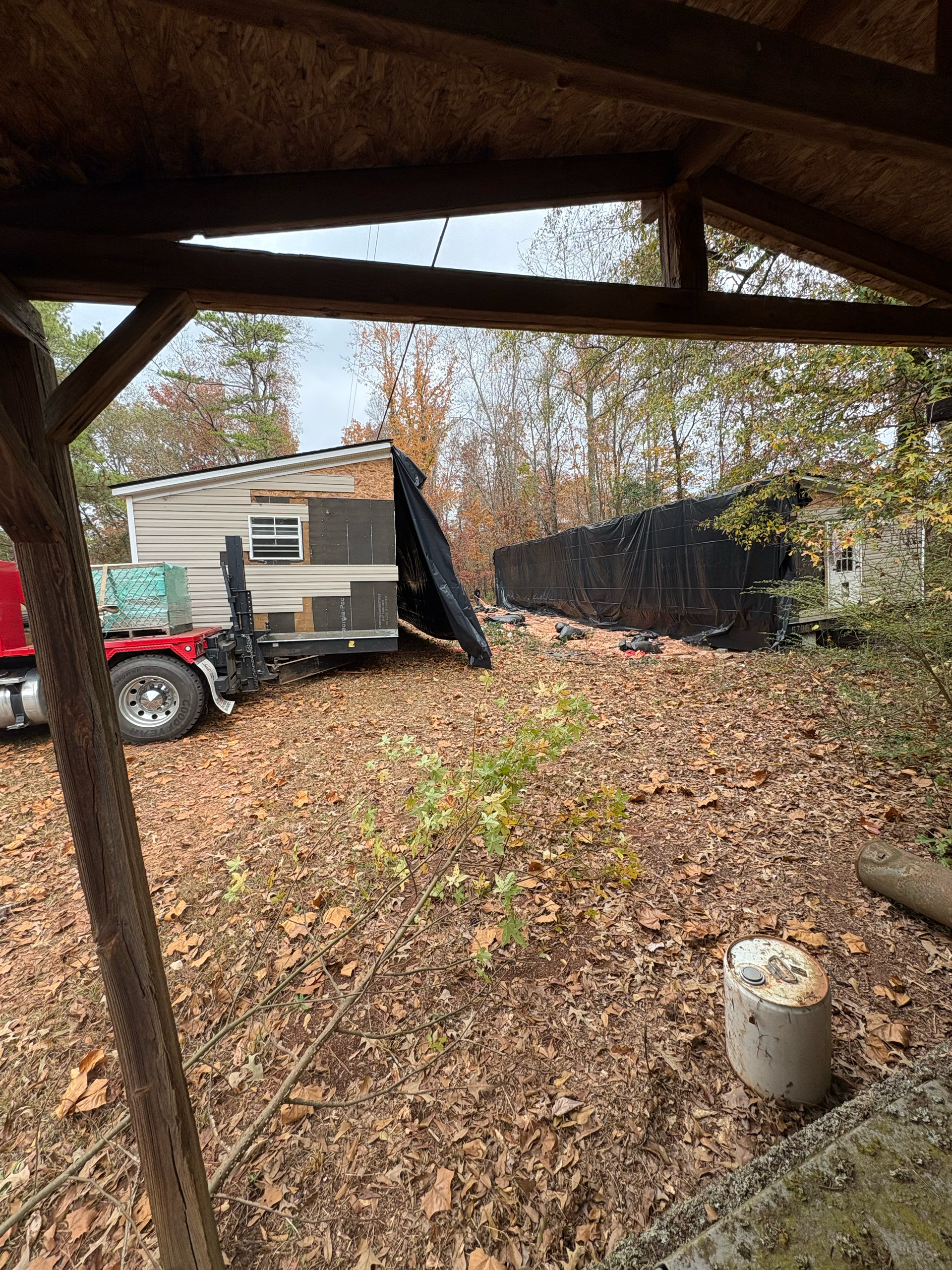 A small, damaged travel trailer parked in a wooded area under a shed, viewed from a shaded vantage point.