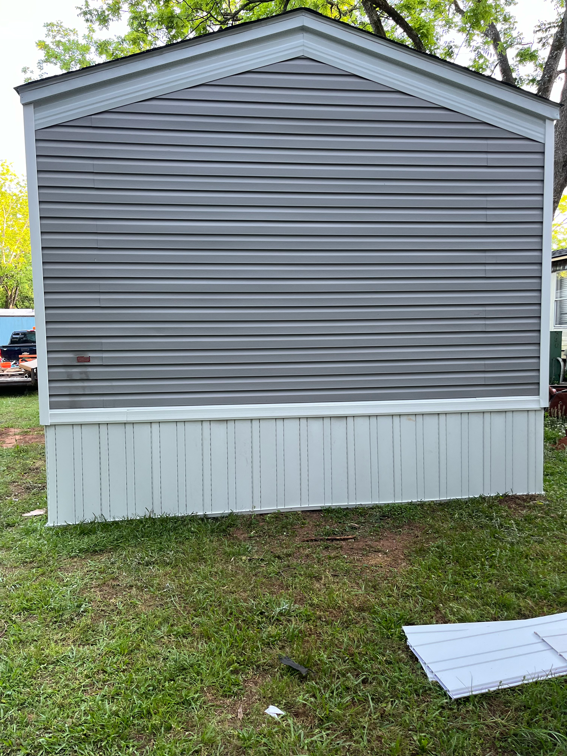 Gray vinyl siding on the wall of a mobile home with white skirting above a green lawn.