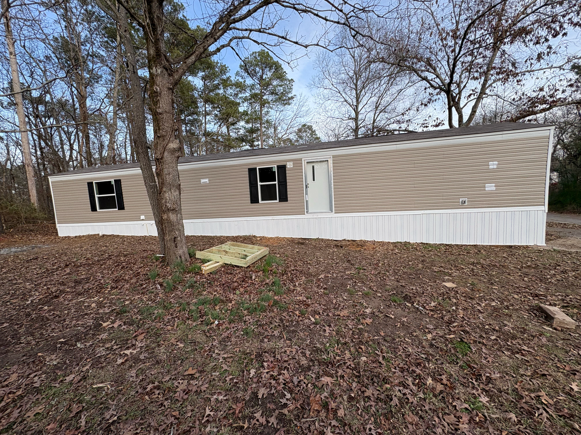 A tan mobile home with white skirting sits on a leaf-covered lot surrounded by trees under a blue sky.
