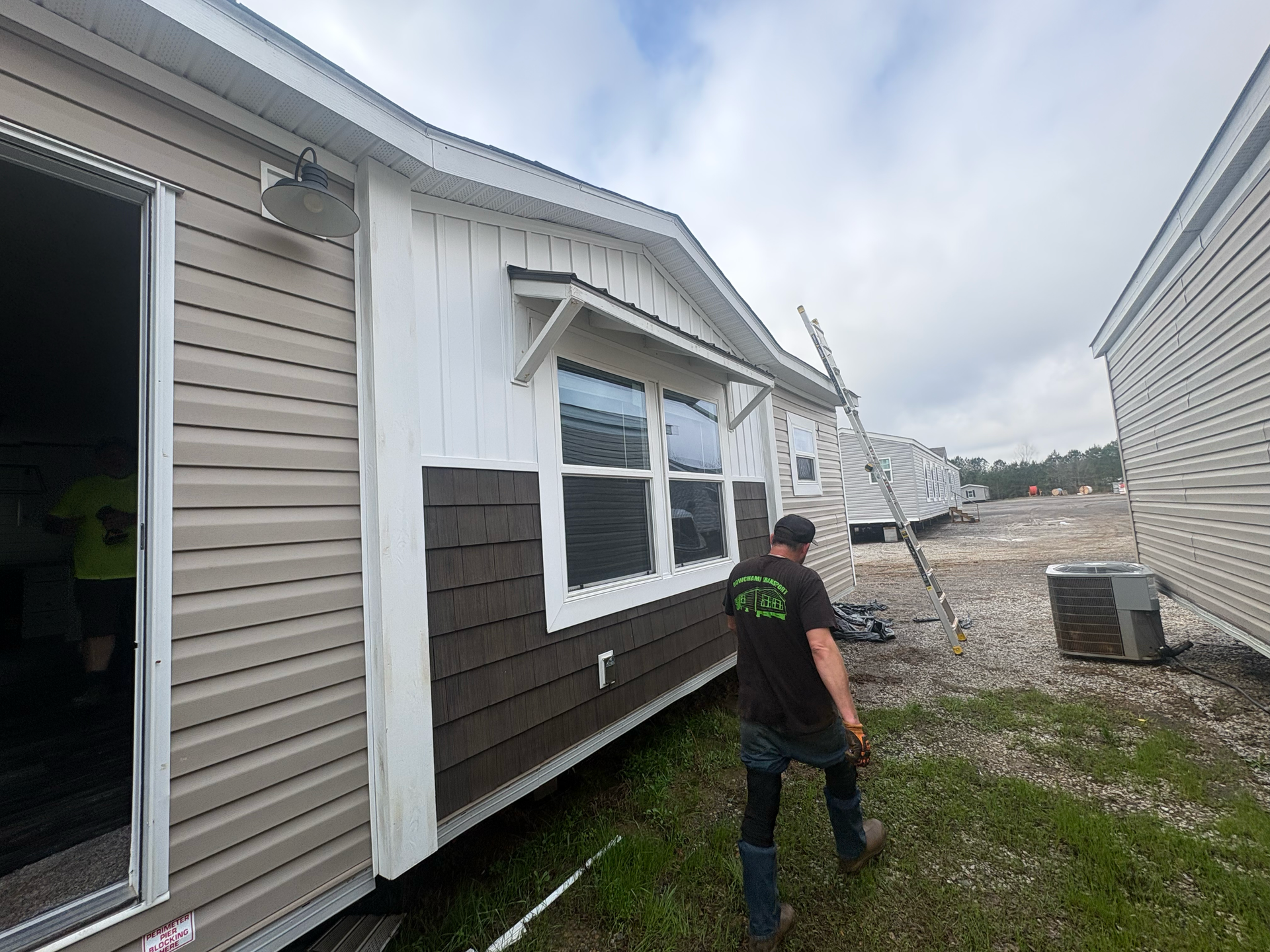 A worker walks past the side of a manufactured home featuring tan horizontal siding, white trim, and brown shingles.
