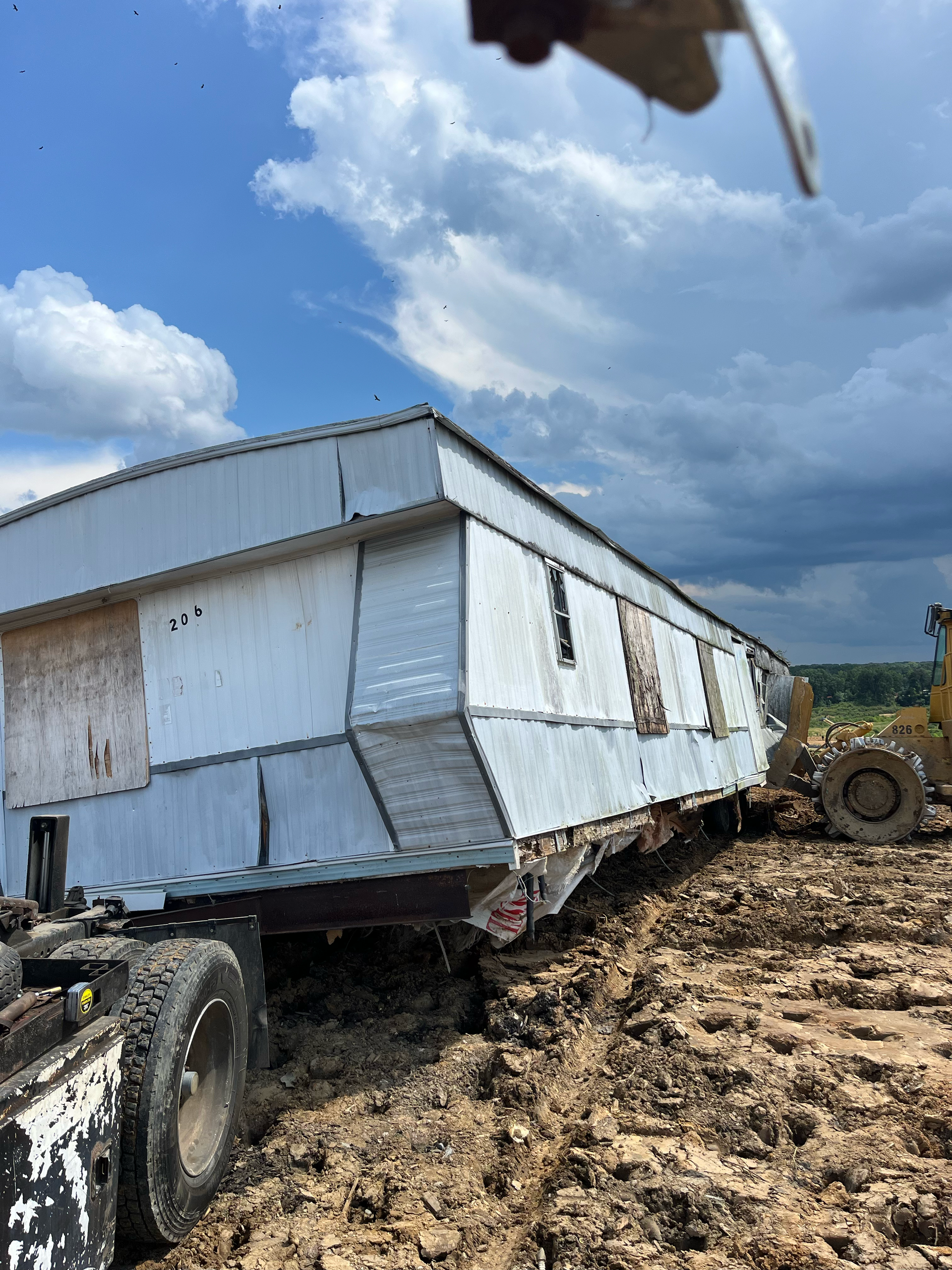 A white mobile home being transported through deep, muddy terrain by a heavy-duty vehicle under a cloudy sky.