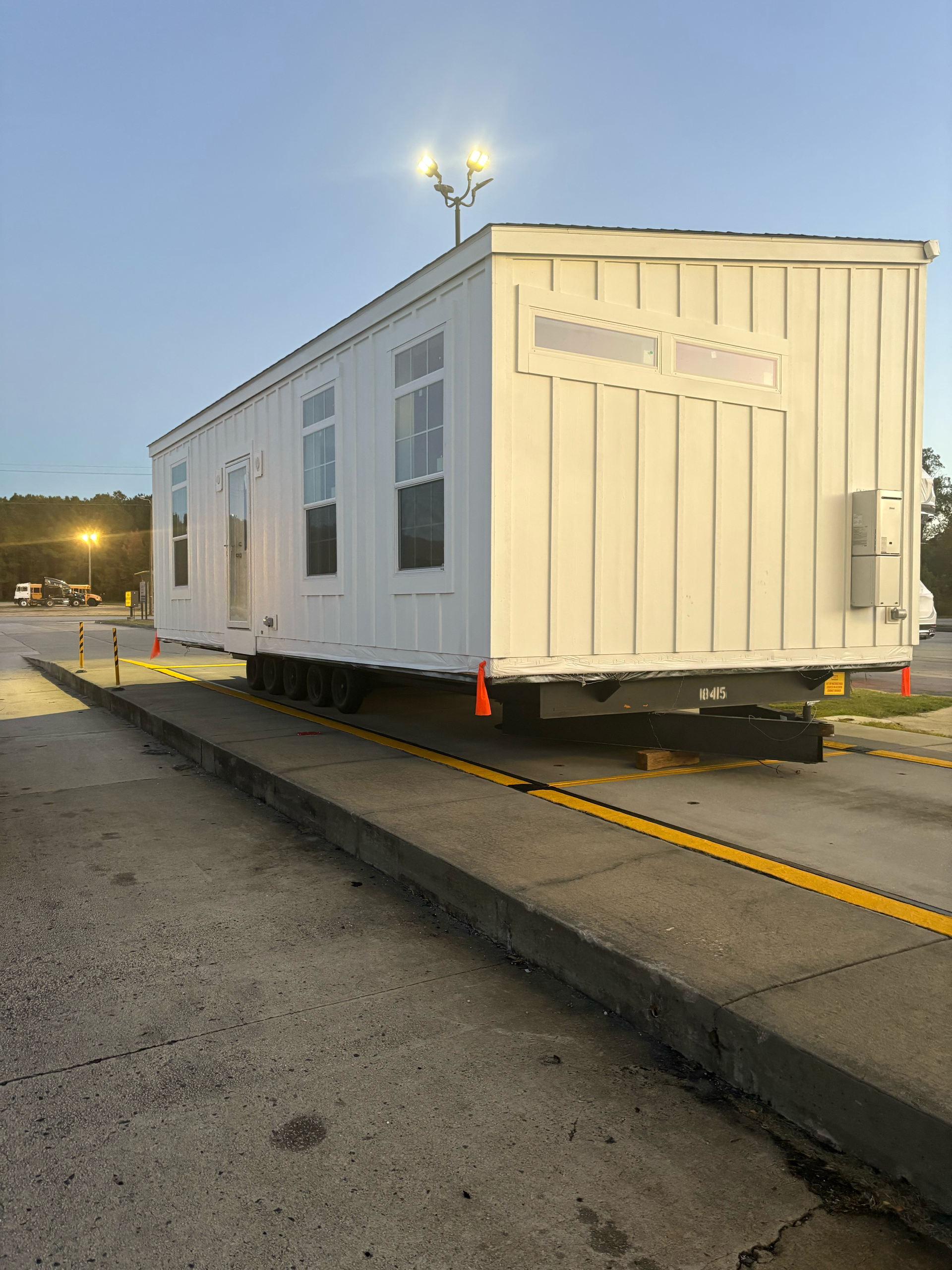 A white modular building on a trailer chassis, positioned on a concrete pad under outdoor lighting at dusk.