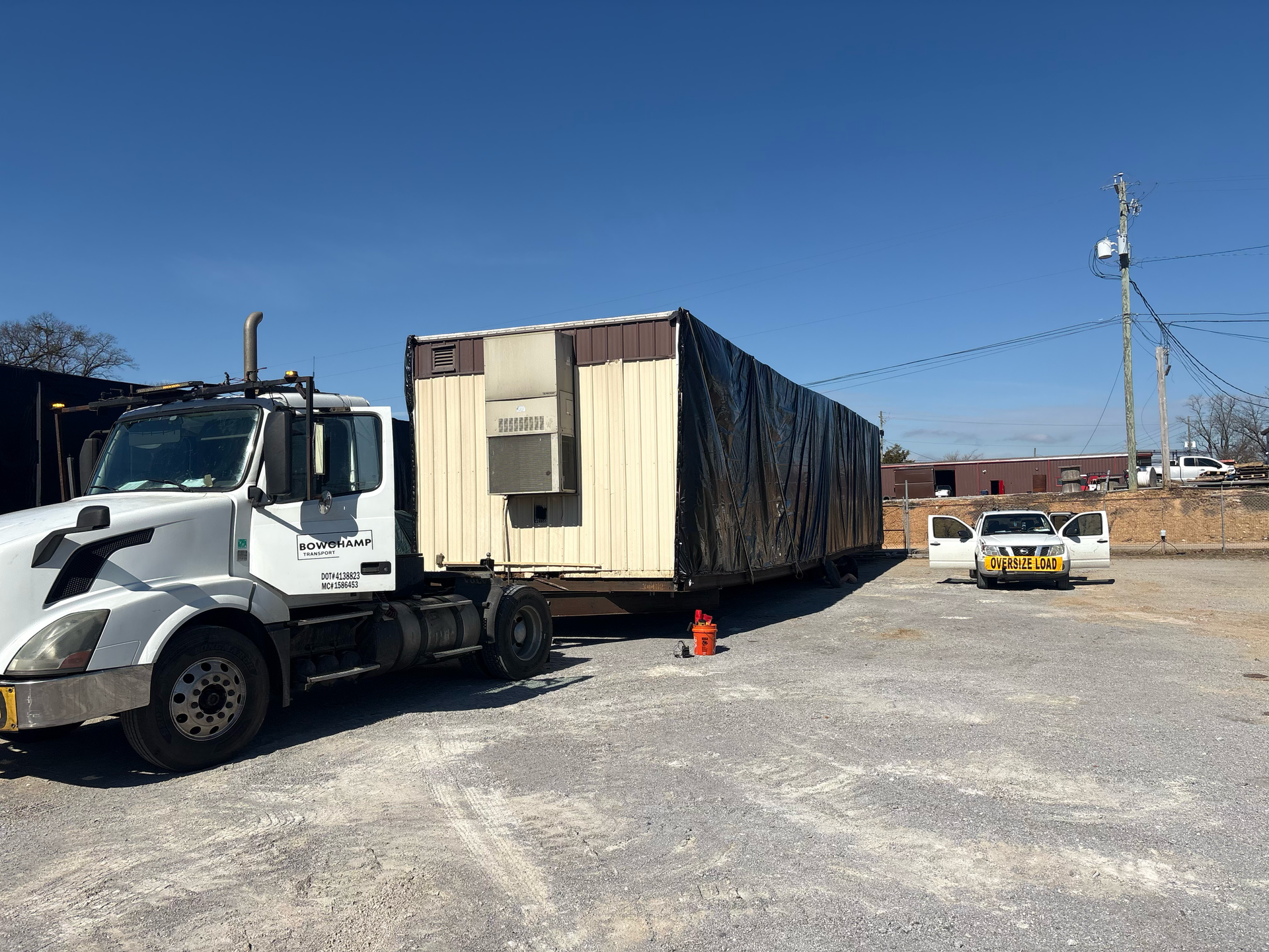 A white semi-truck hauls a large, beige and black mobile structure across a gravel lot under a clear blue sky.