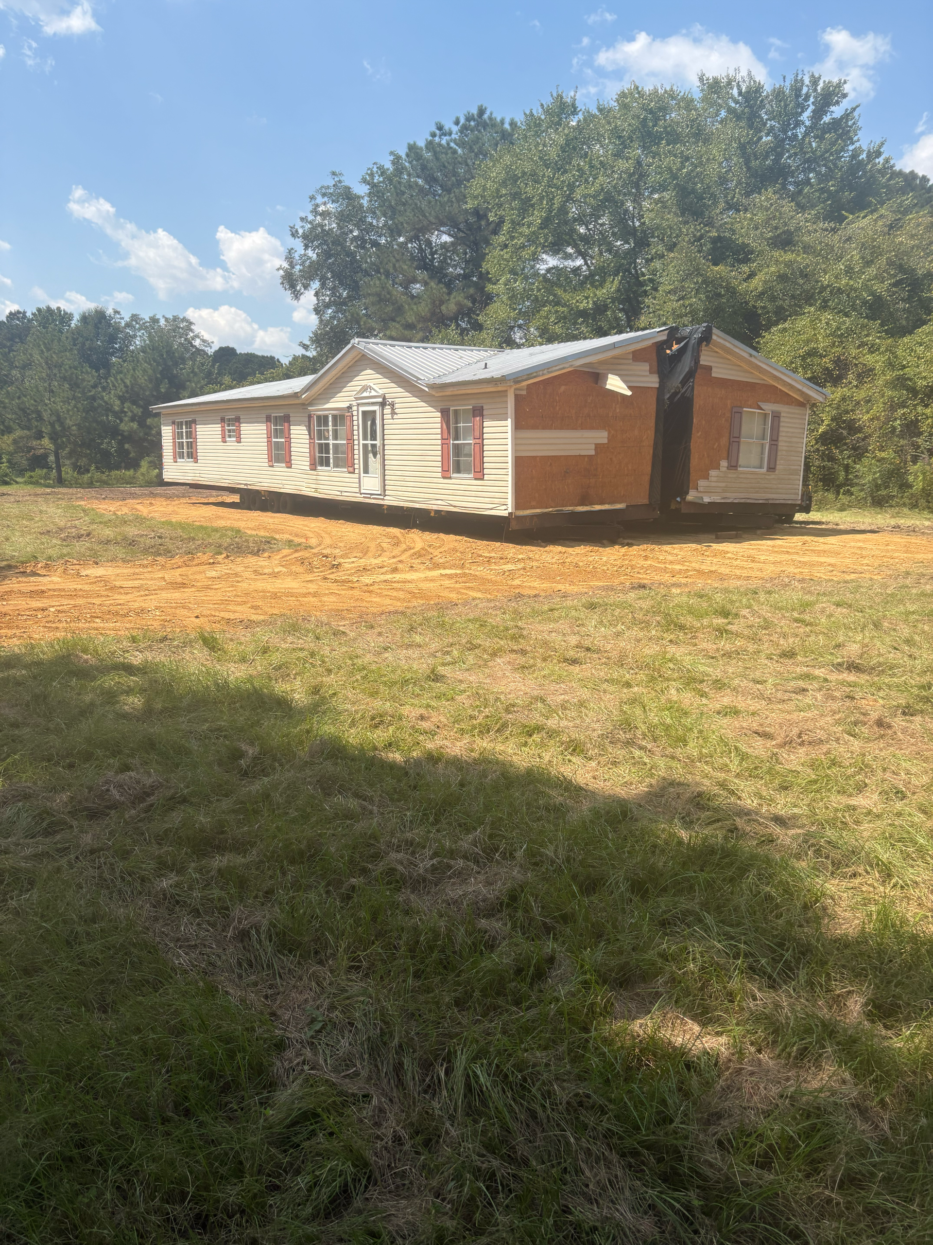 A manufactured home sits in a grassy field on a patch of bare, orange dirt under a bright blue sky.