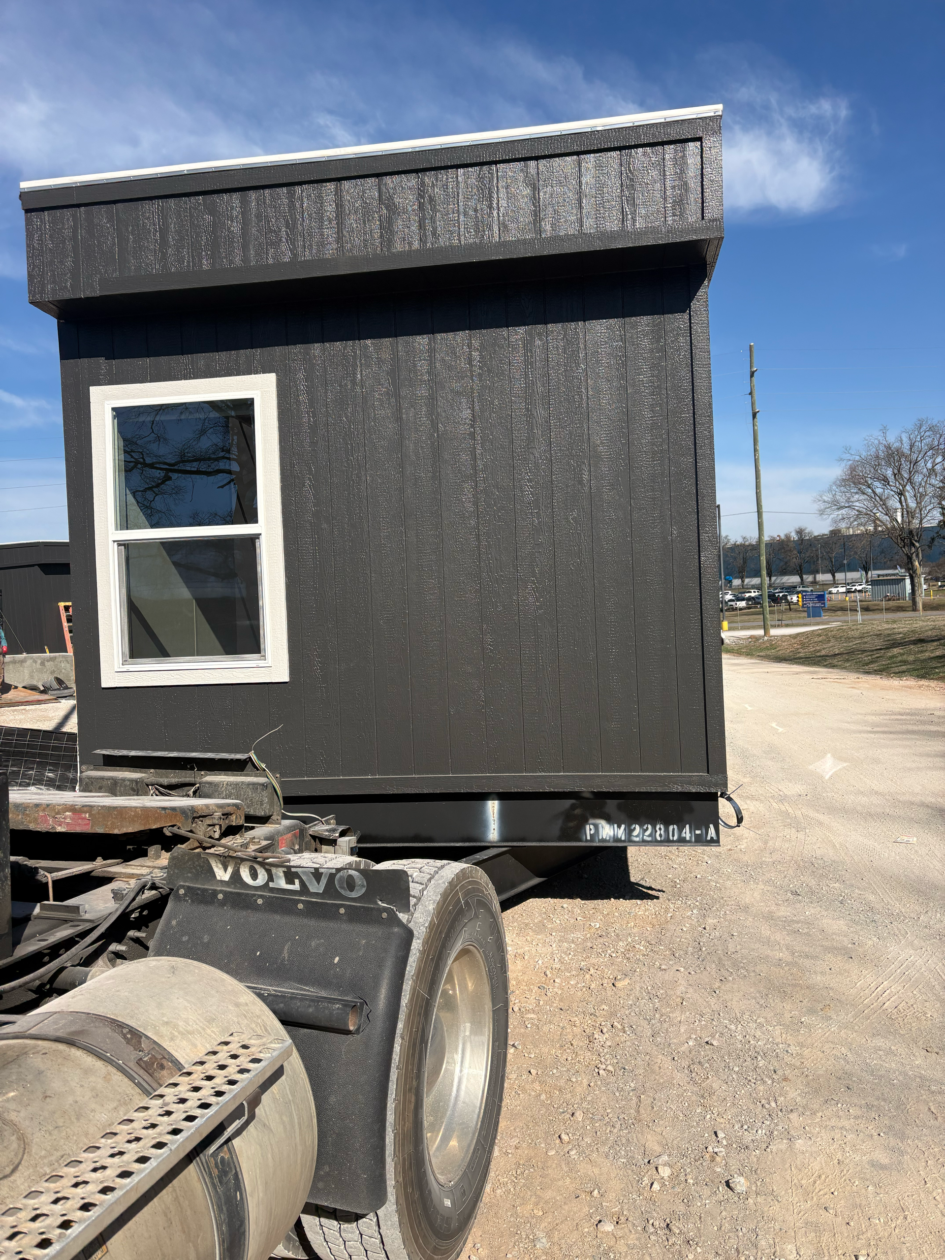 A black, boxy, windowed modular structure secured to the back of a large semi-truck on a gravel lot under a blue sky.
