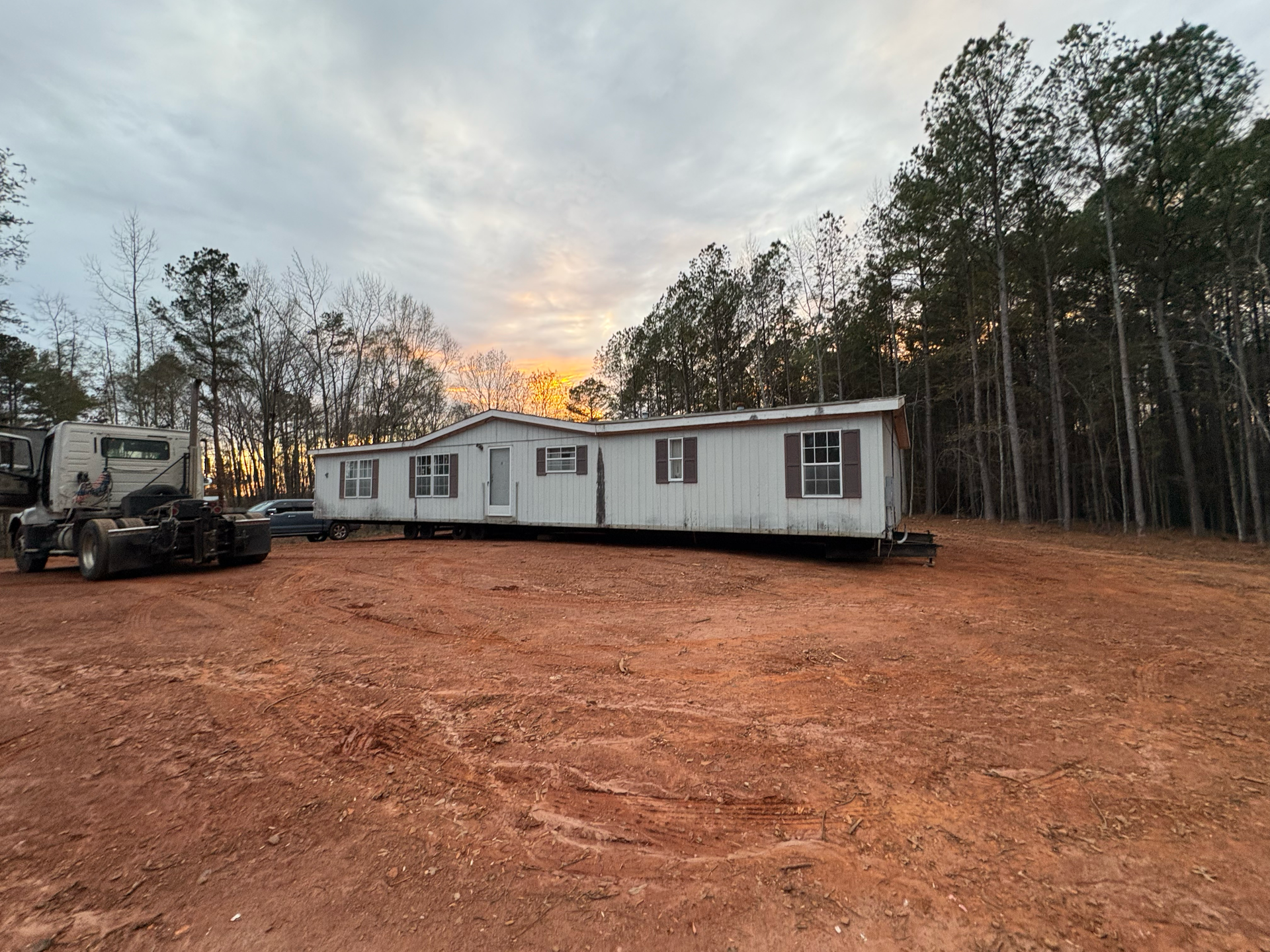 A light-colored mobile home being towed across a red-dirt lot by a truck at dusk near a treeline.