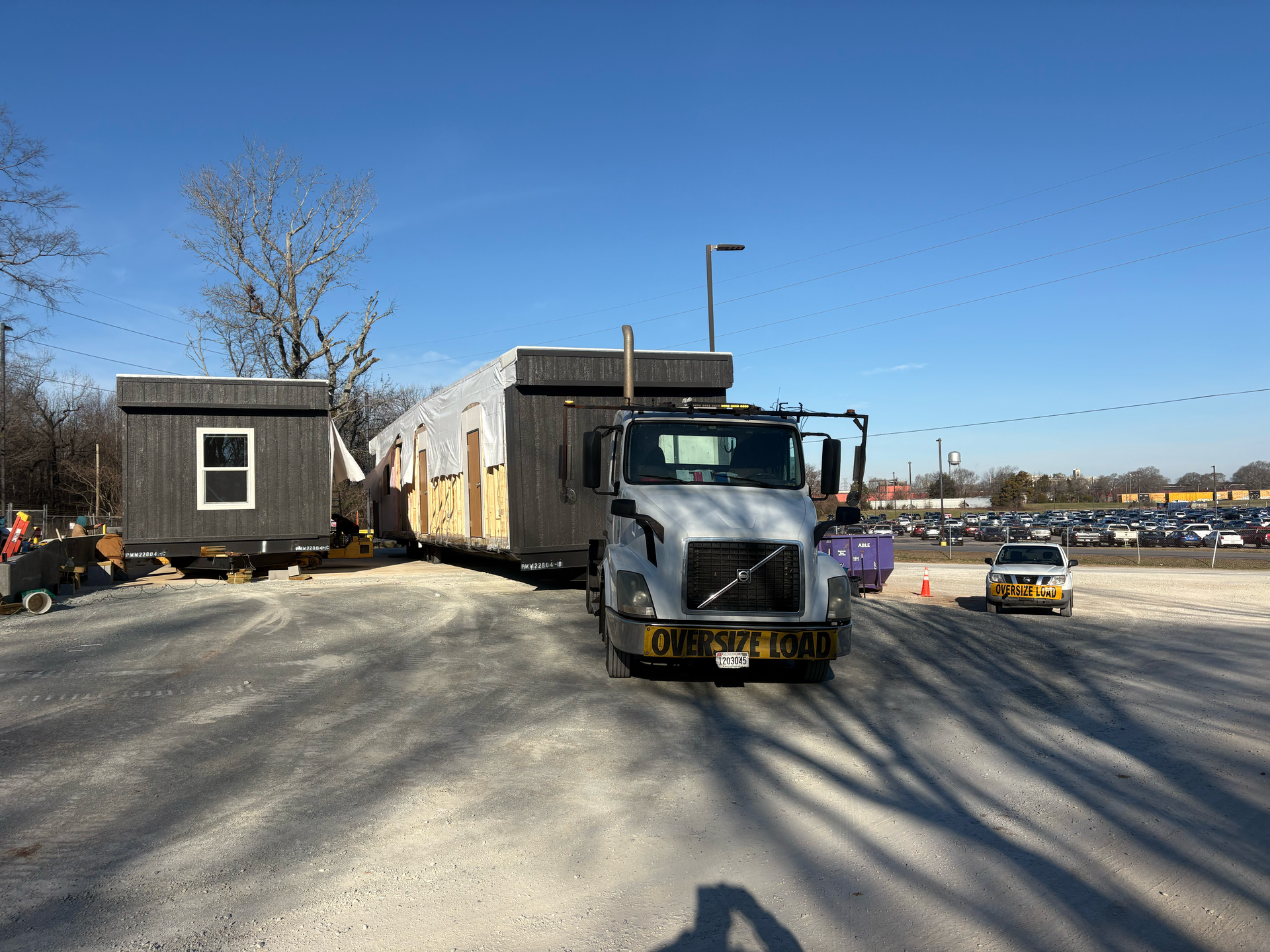 A white Volvo semi-truck hauls a modular home section across a gravel lot on a sunny day.