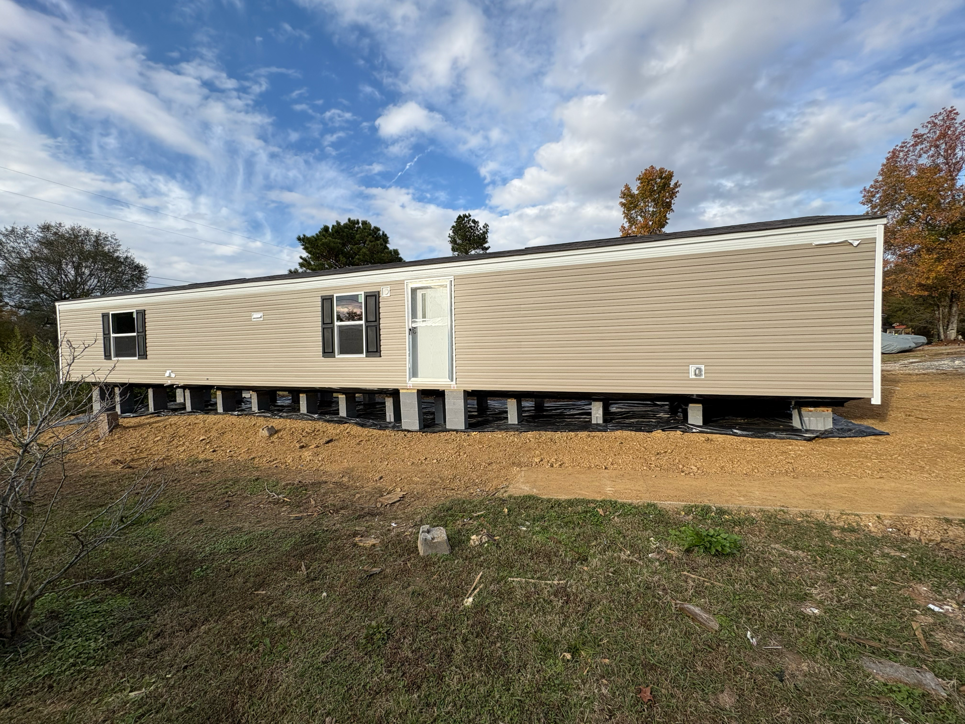A tan mobile home sits on a foundation of concrete blocks in a field under a cloudy blue sky.