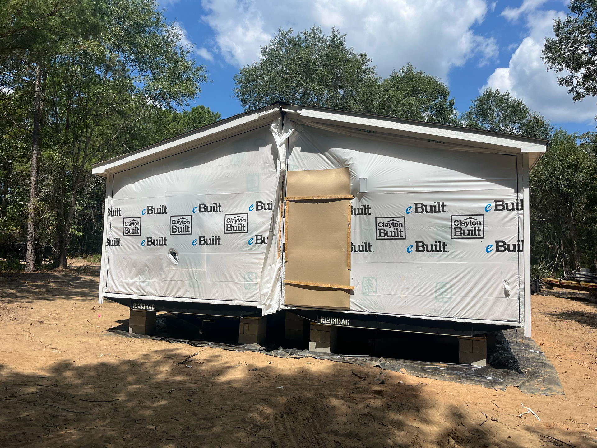 A small, unfinished house frame on concrete blocks in a wooded area, covered in white building wrap.