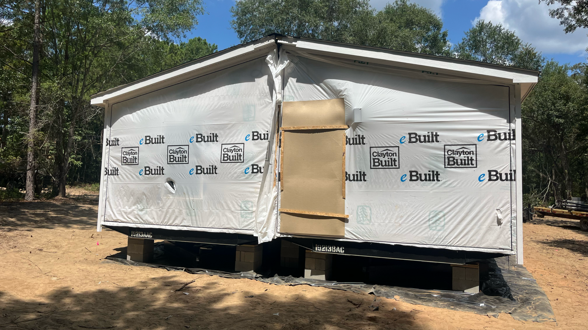 White portable building under construction with wrapped exterior, front door panel, and wooded dirt site.